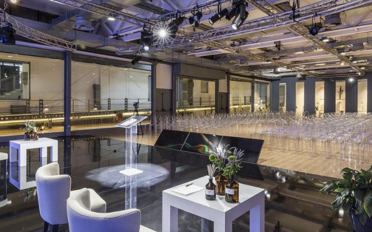 Una sala di conferenze vuota con sedie trasparenti e un palco con decorazioni floreali, luci e apparecchiature di scena, apparentemente pronta per un evento o una cerimonia.