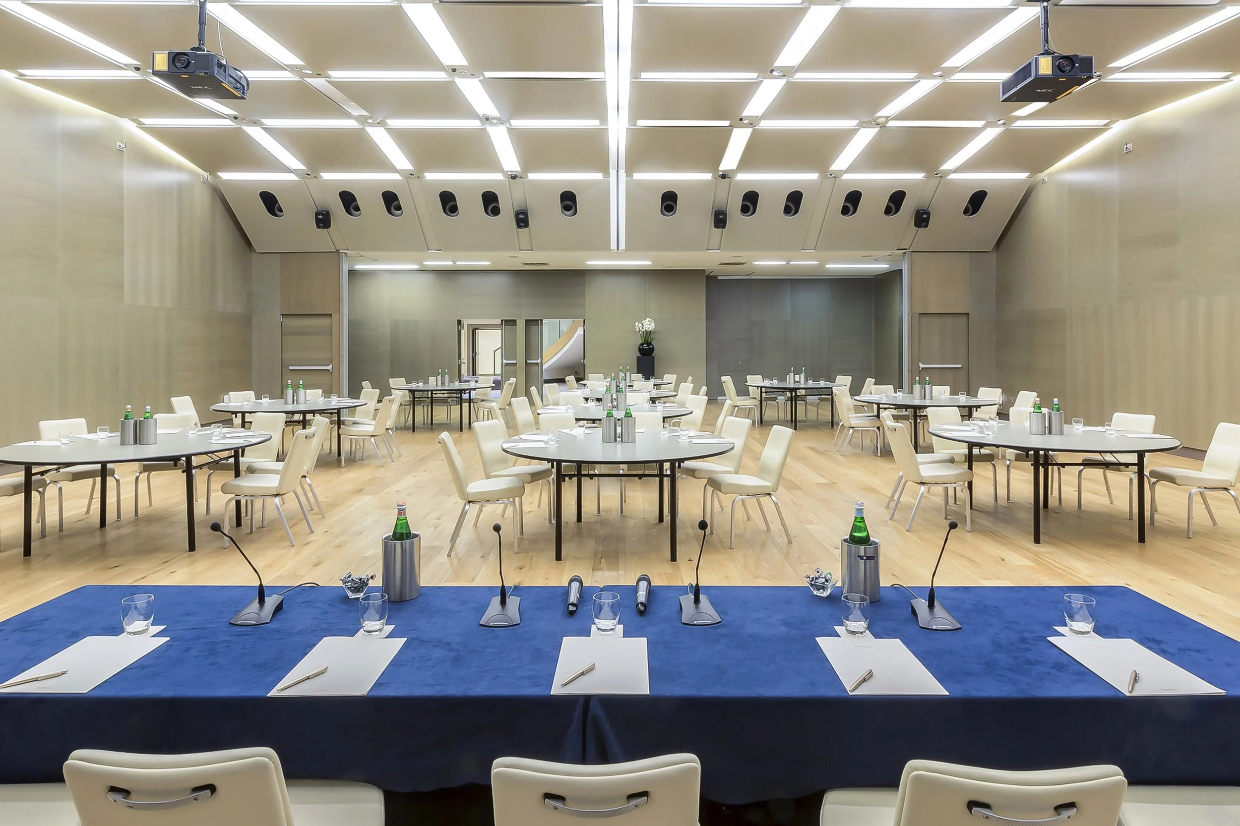 Sala conferenze con tavoli rotondi e sedie bianche, al centro un tavolo con microfoni, bottiglie d'acqua e bicchieri, pareti chiare e soffitto con luci a LED, proiettore sospeso.