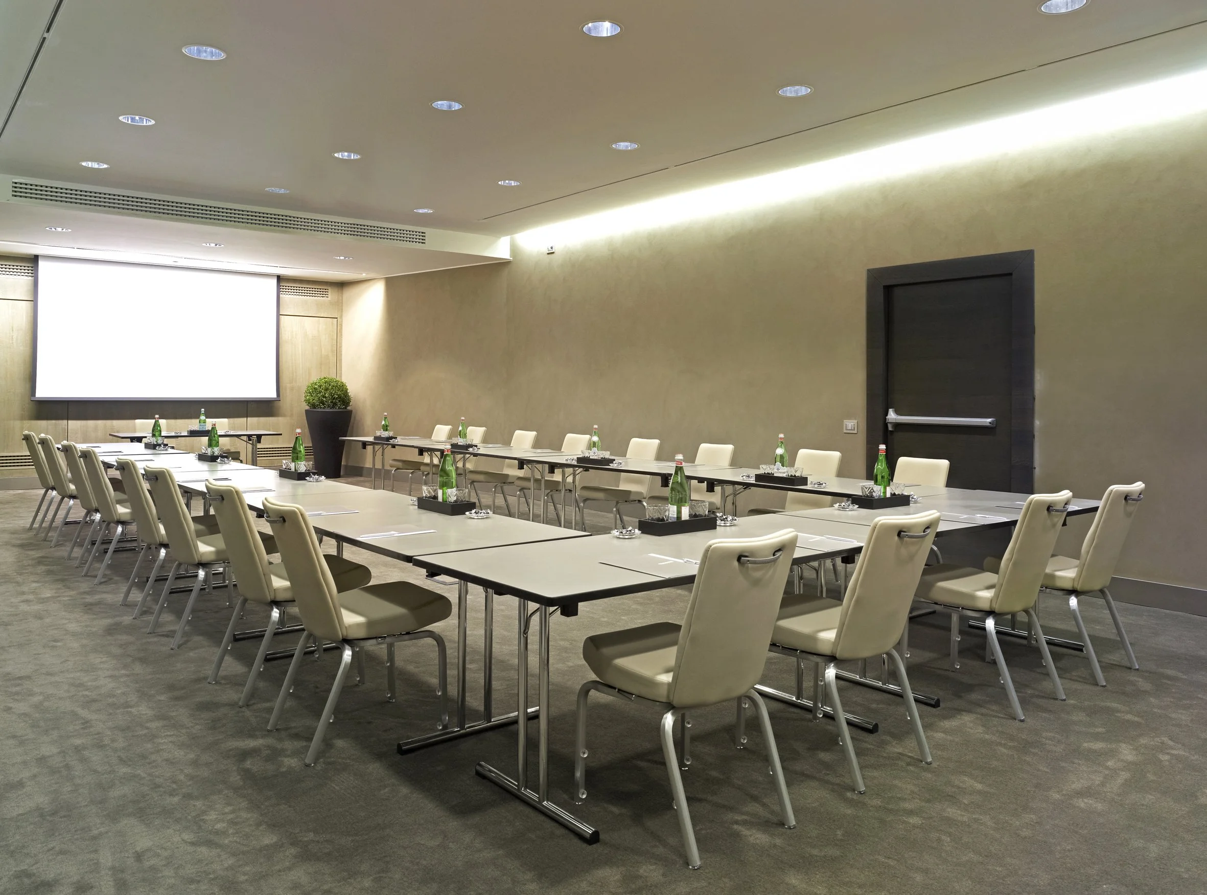 Sala riunioni moderna con tavoli disposti a forma di U, sedie beige, bottiglie d'acqua e bicchieri su ogni tavolo, grande schermo sullo sfondo e piante decorative.