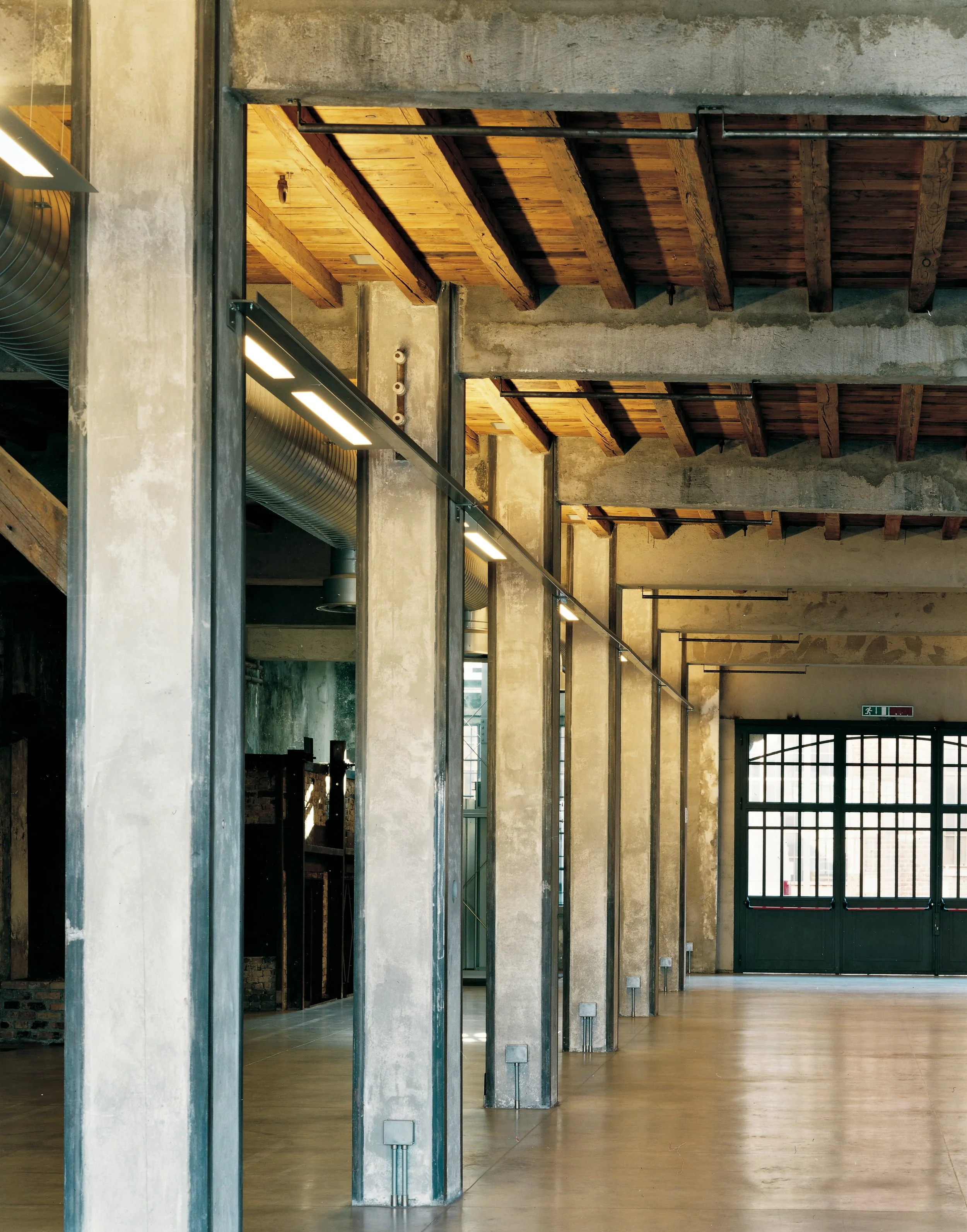 Interno di un edificio industrial con pareti in cemento, soffitto in legno e colonne in metallo, con ampio spazio e grandi porte di uscita.