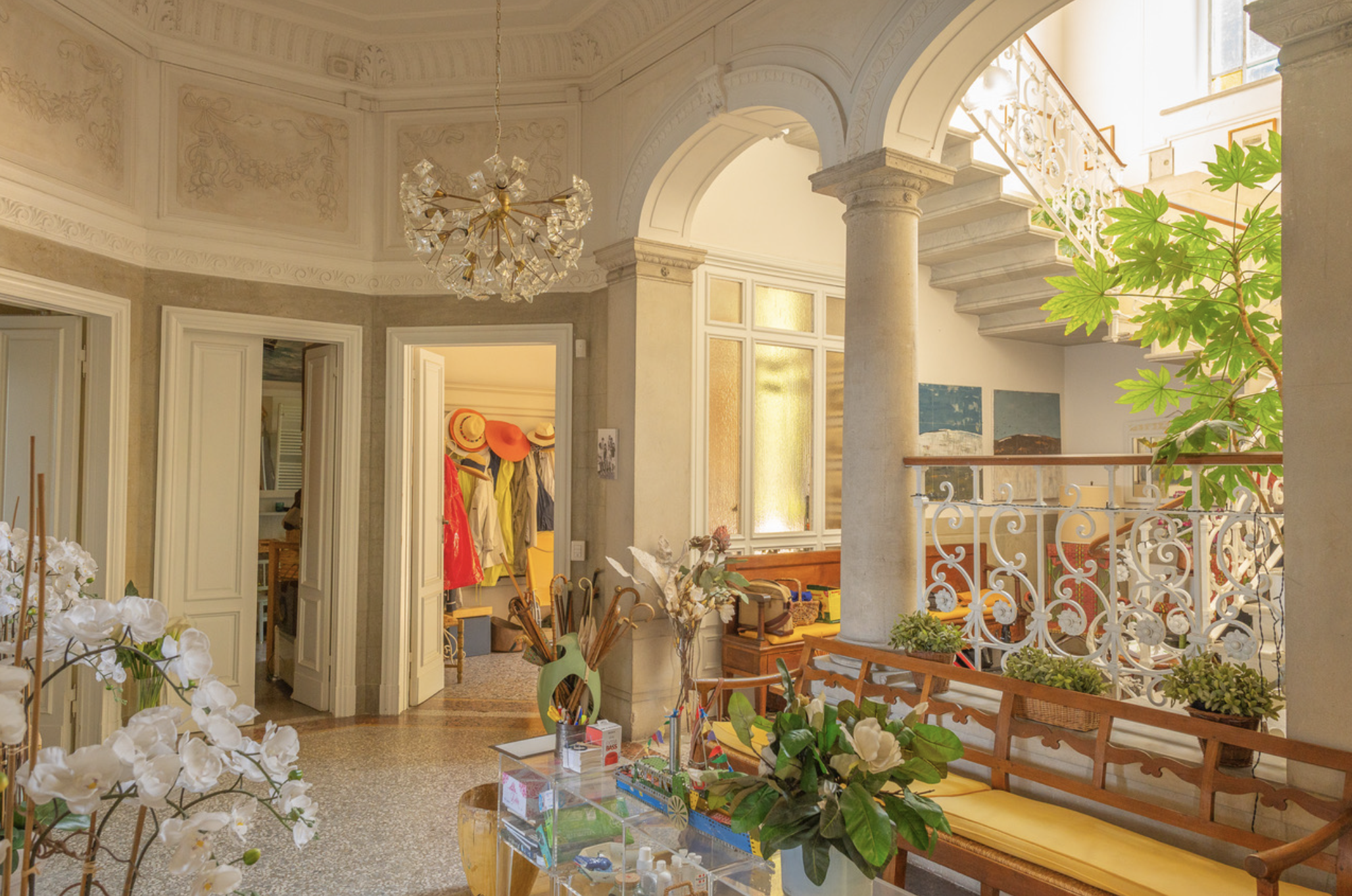 Interno di una casa con poltrona, panchina con cuscini gialli, balcone con ringhiera decorativa, pianta verde, arco, lampadario di cristallo e ingresso a una stanza con cappelli e vestiti appesi.