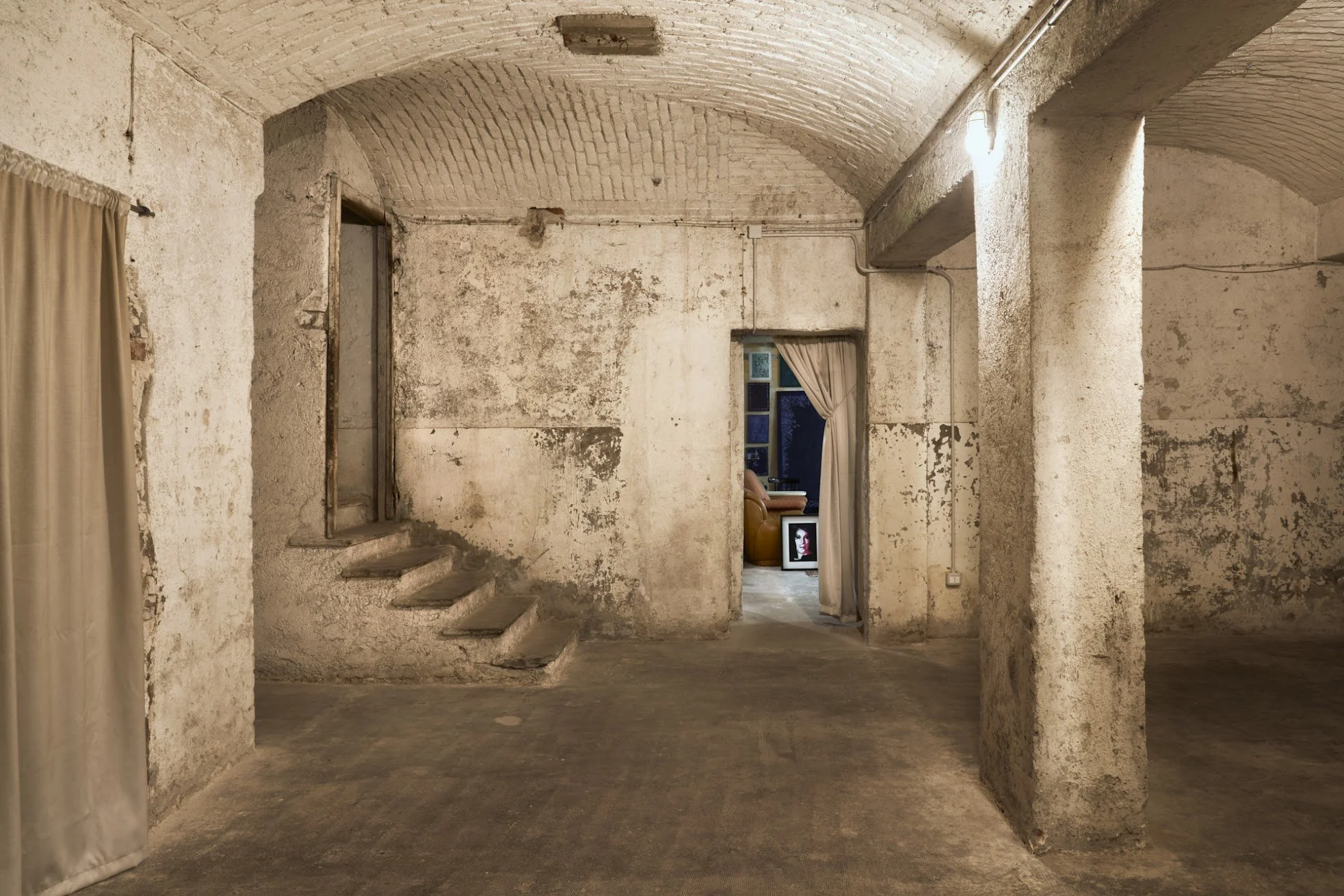 Interno di una stanza con pareti e soffitto in pietra, arredo minimo, presenza di un quadro di fronte.