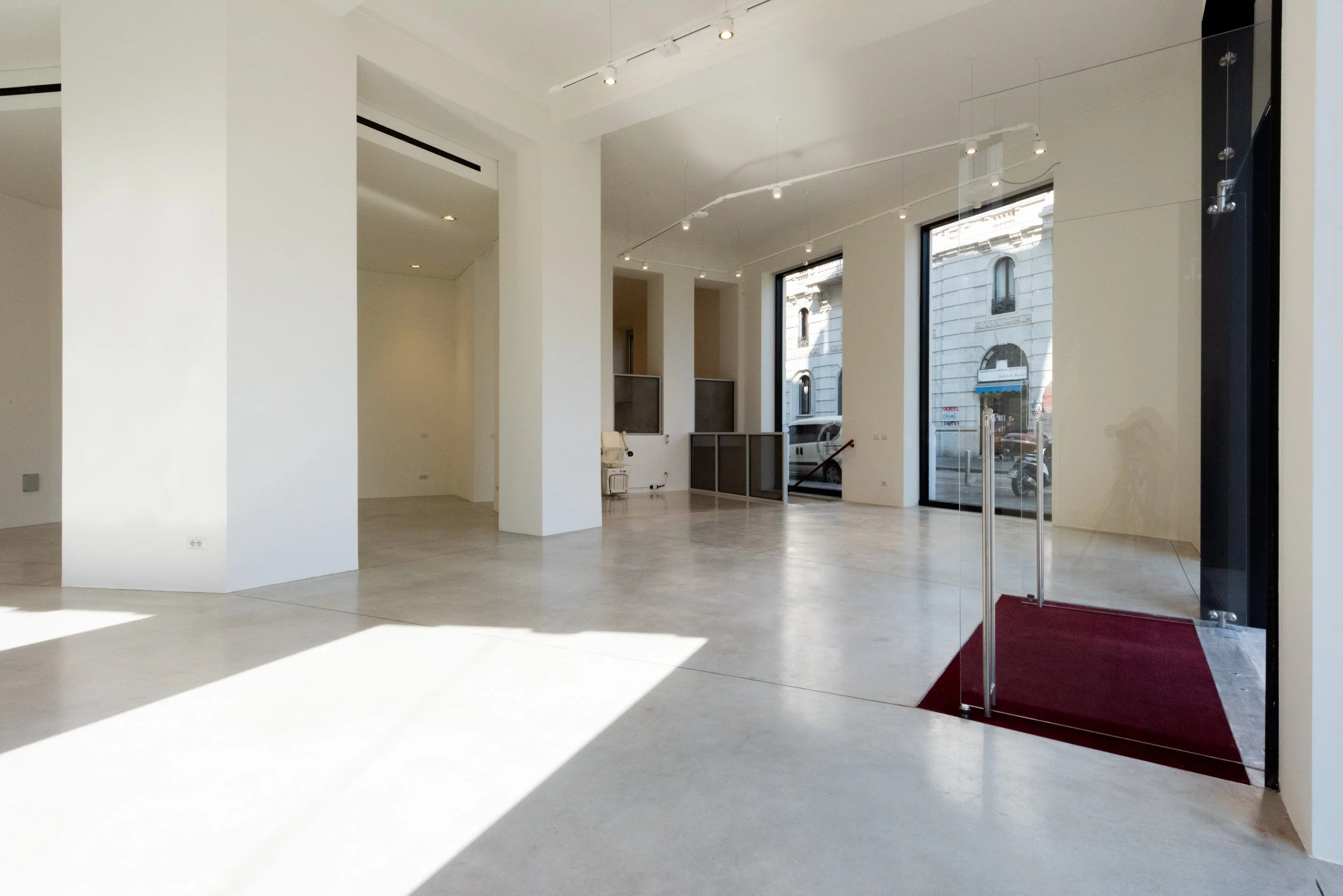 Interno di un edificio con pareti bianche, grandi finestre e pavimento in calcestruzzo. L'ingresso ha un tappeto rosso e grandi porte di vetro.