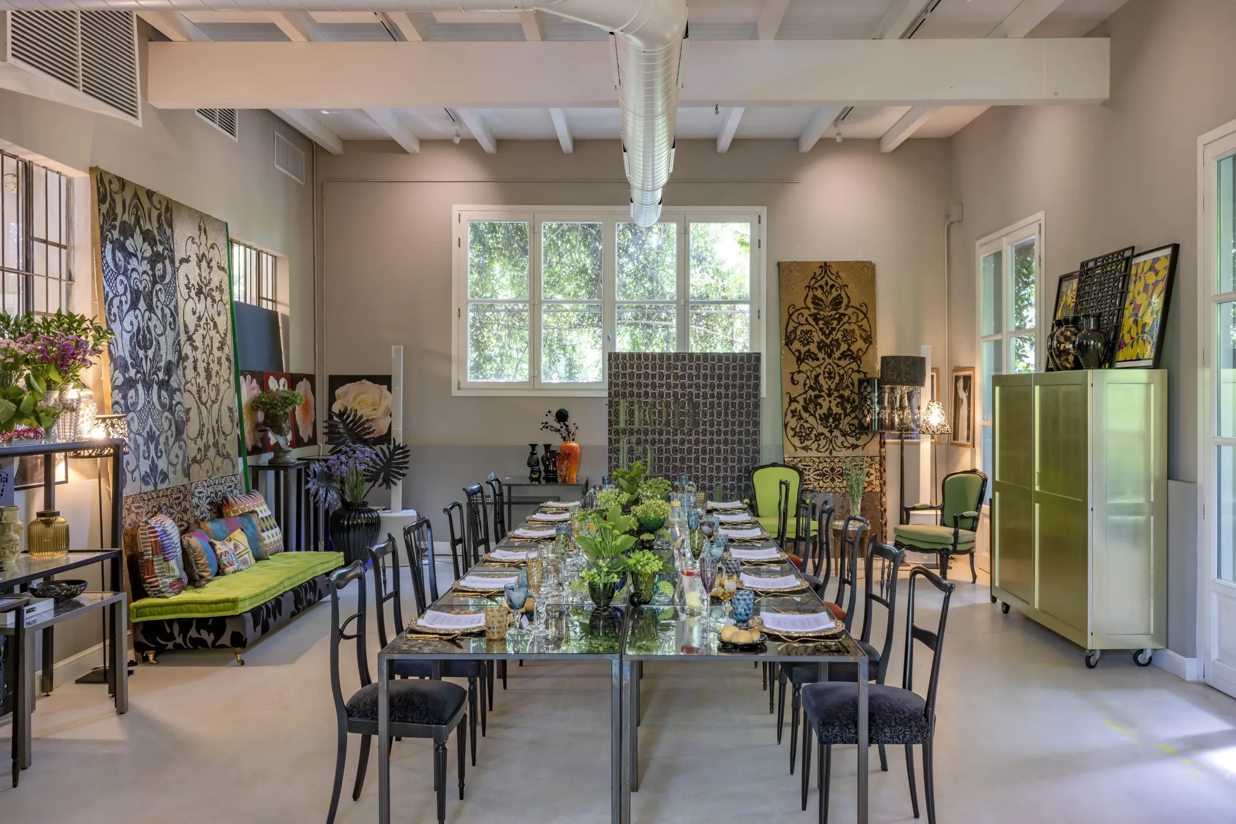 Sala da pranzo elegante con tavolo lungo, coperto con tovaglioli e decorazioni floreali, sedie nere e divano con cuscini colorati, pareti decorate con tende e quadri, illuminata da grandi finestre.