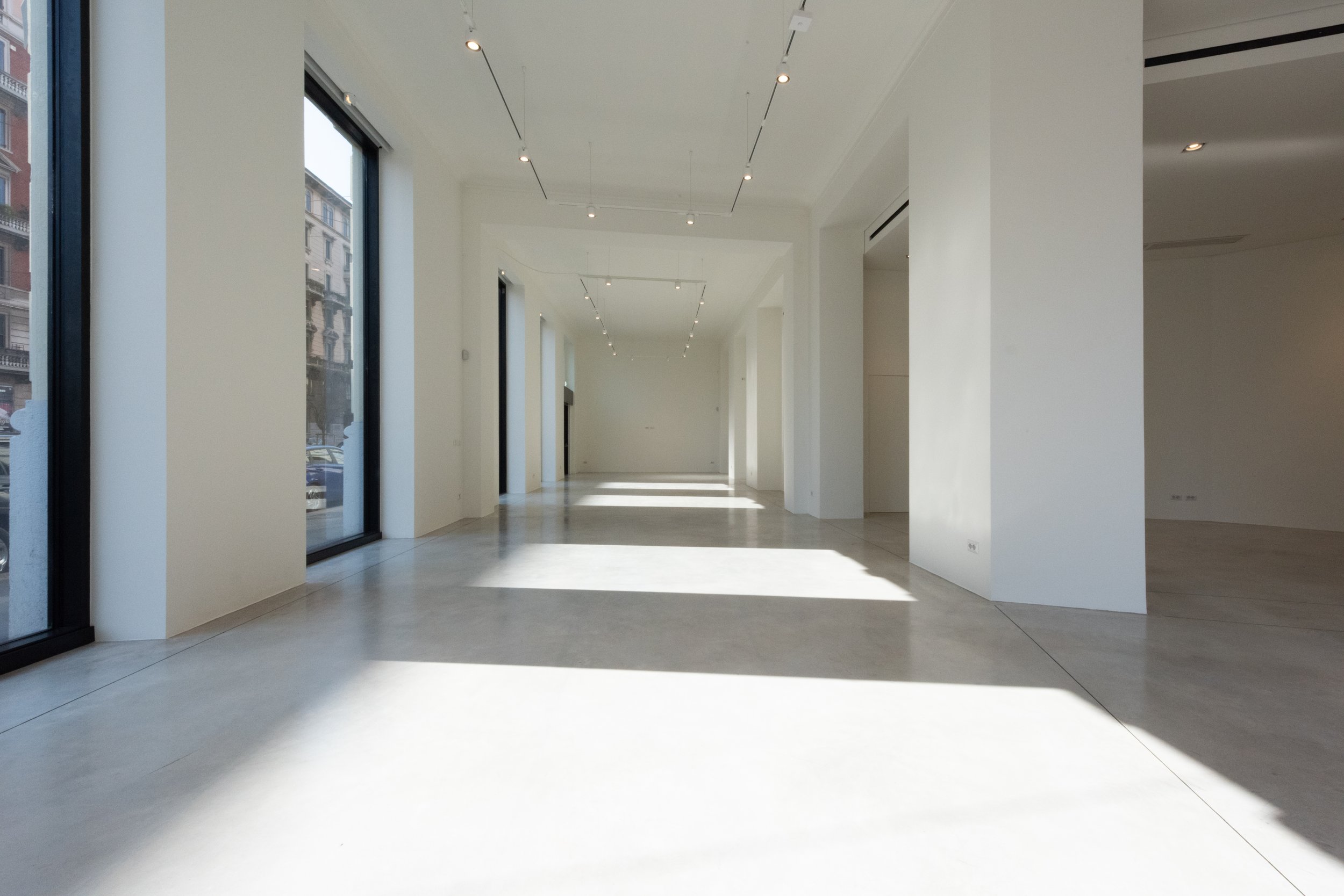 Interno di un edificio moderno con pareti bianche, grandi finestre e pavimento in piastrelle chiara, illuminato dalla luce naturale.