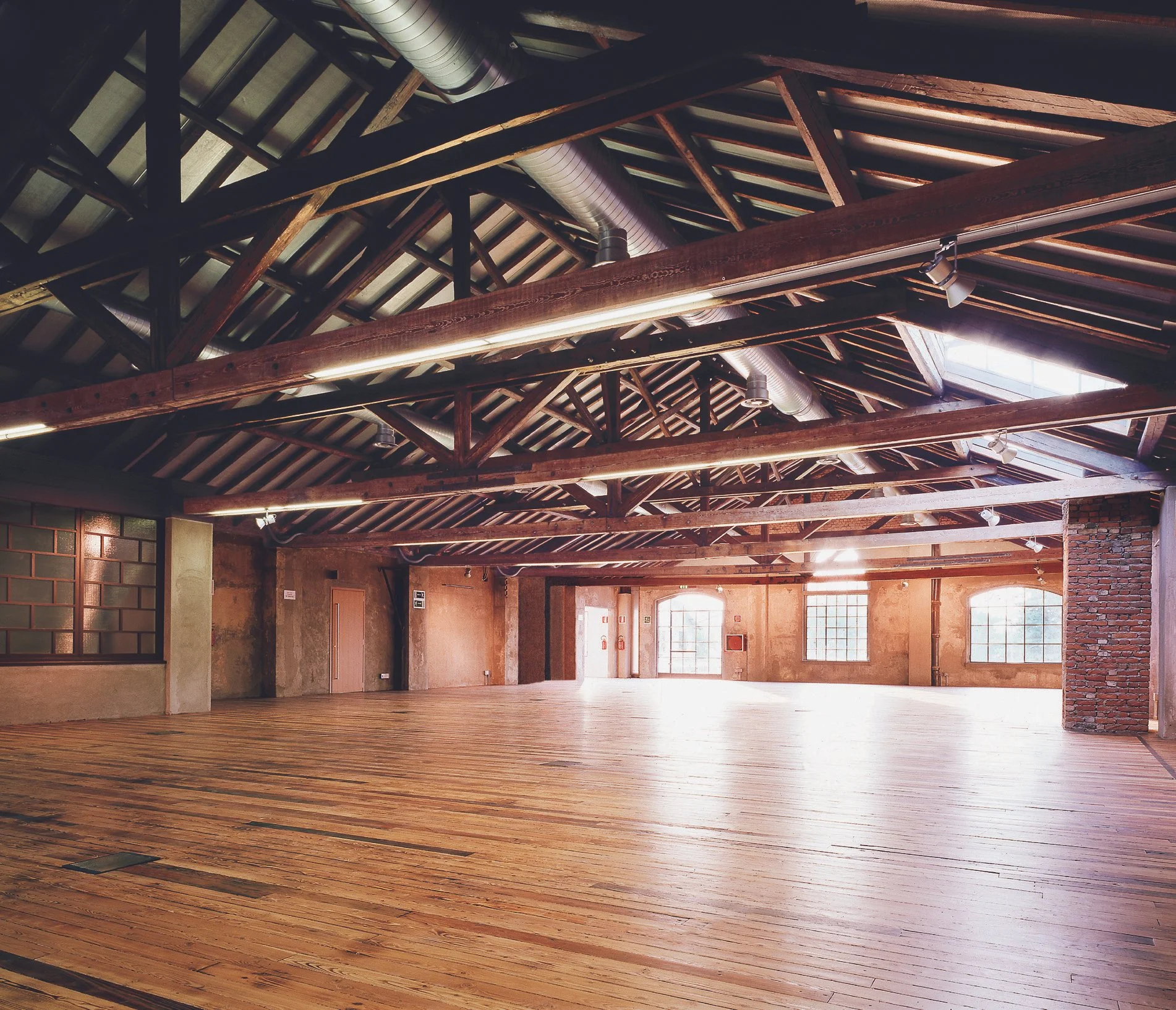Interno di un grande spazio aperto con soffitto in legno e grandi finestre, pavimento in legno e pareti in mattoni e cemento