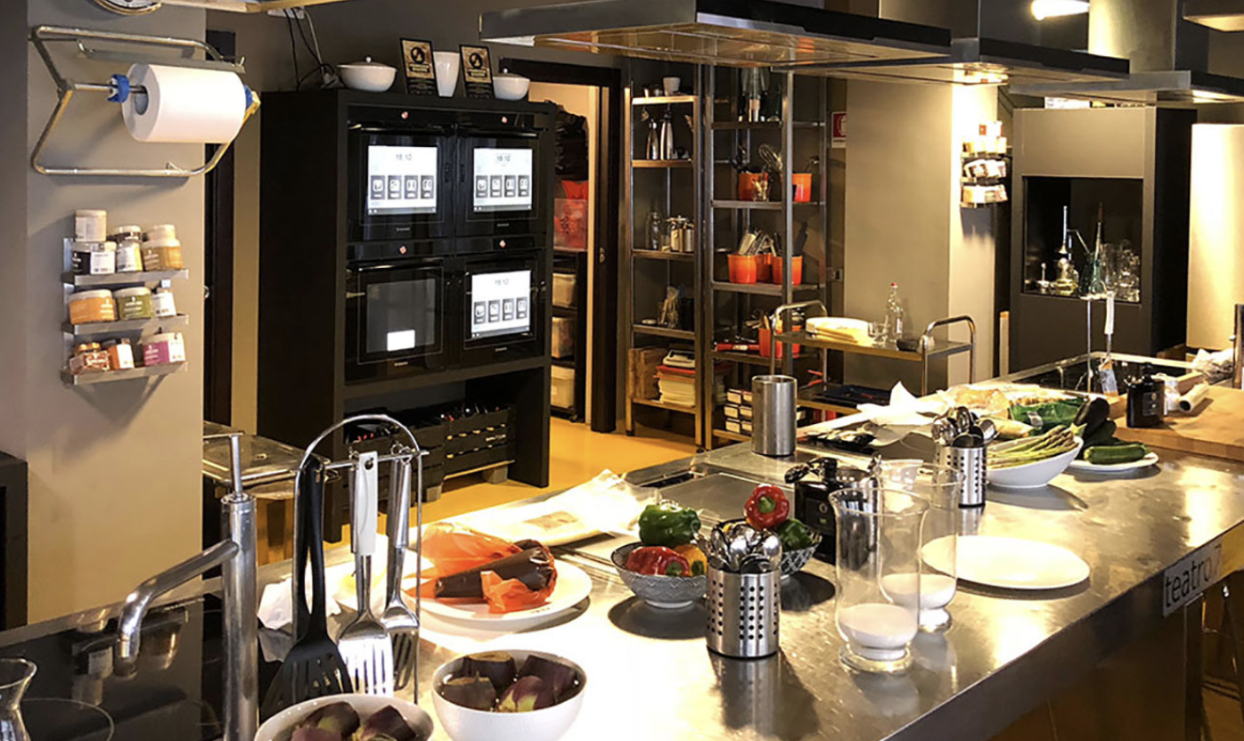 Una cucina moderna con un banco di lavoro in acciaio, utensili, verdure e attrezzature da cucina disposte sul piano di lavoro, scaffali con utensili e spezie, e schermi digitali in uno spazio ben illuminato.