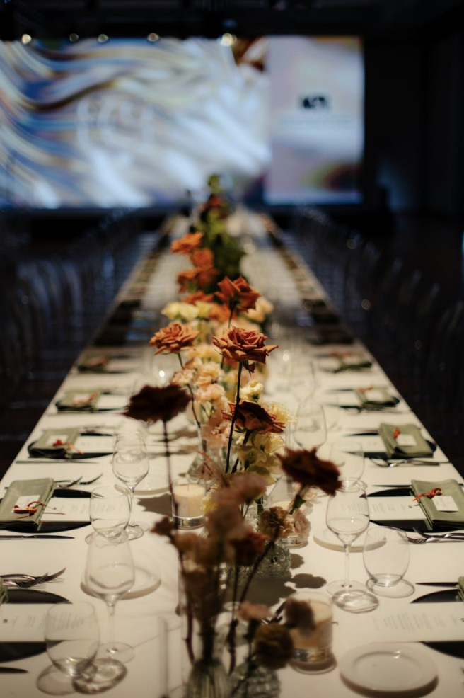 Tavola da pranzo lunga decorata con fiori artificiali. Sul tavolo ci sono bicchieri, piatti, posate e menu. Sfondo con grande schermo in una sala elegante.