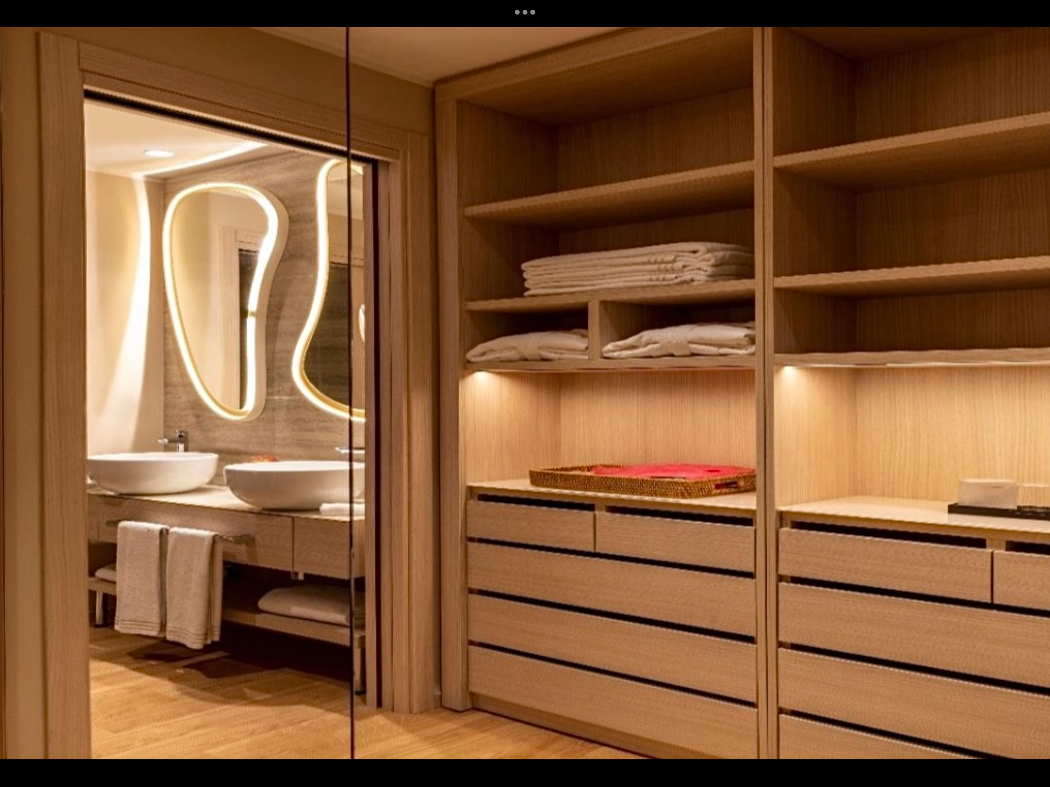 L'interno di una stanza con armadi in legno e una porta in vetro che conduce a un bagno con lavabi tondi, specchi illuminati e asciugamani.