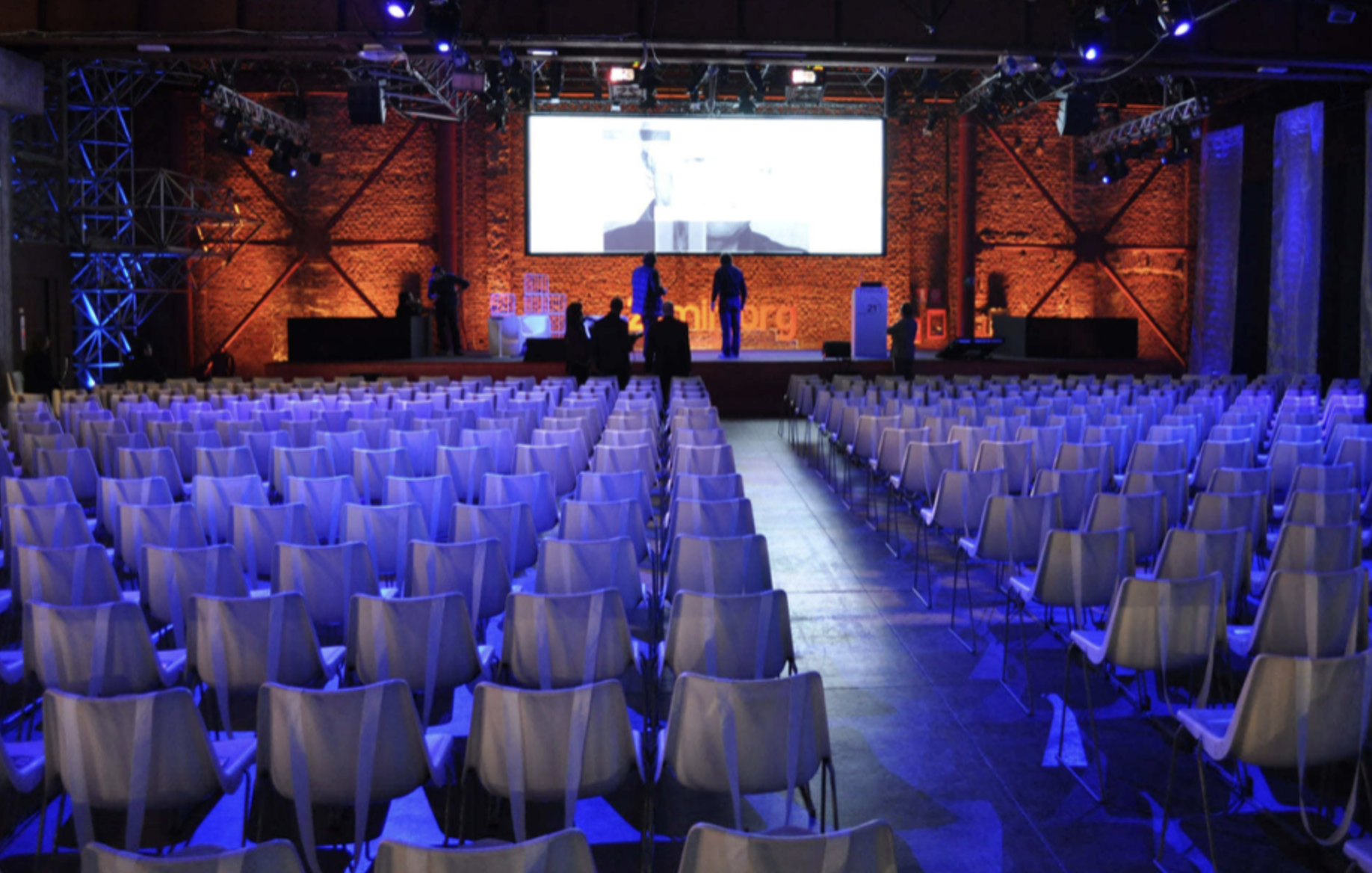 Sala conferenze con molte sedie bianche disposte in file, palco con grande schermo e alcuni persone che si preparano prima di un evento