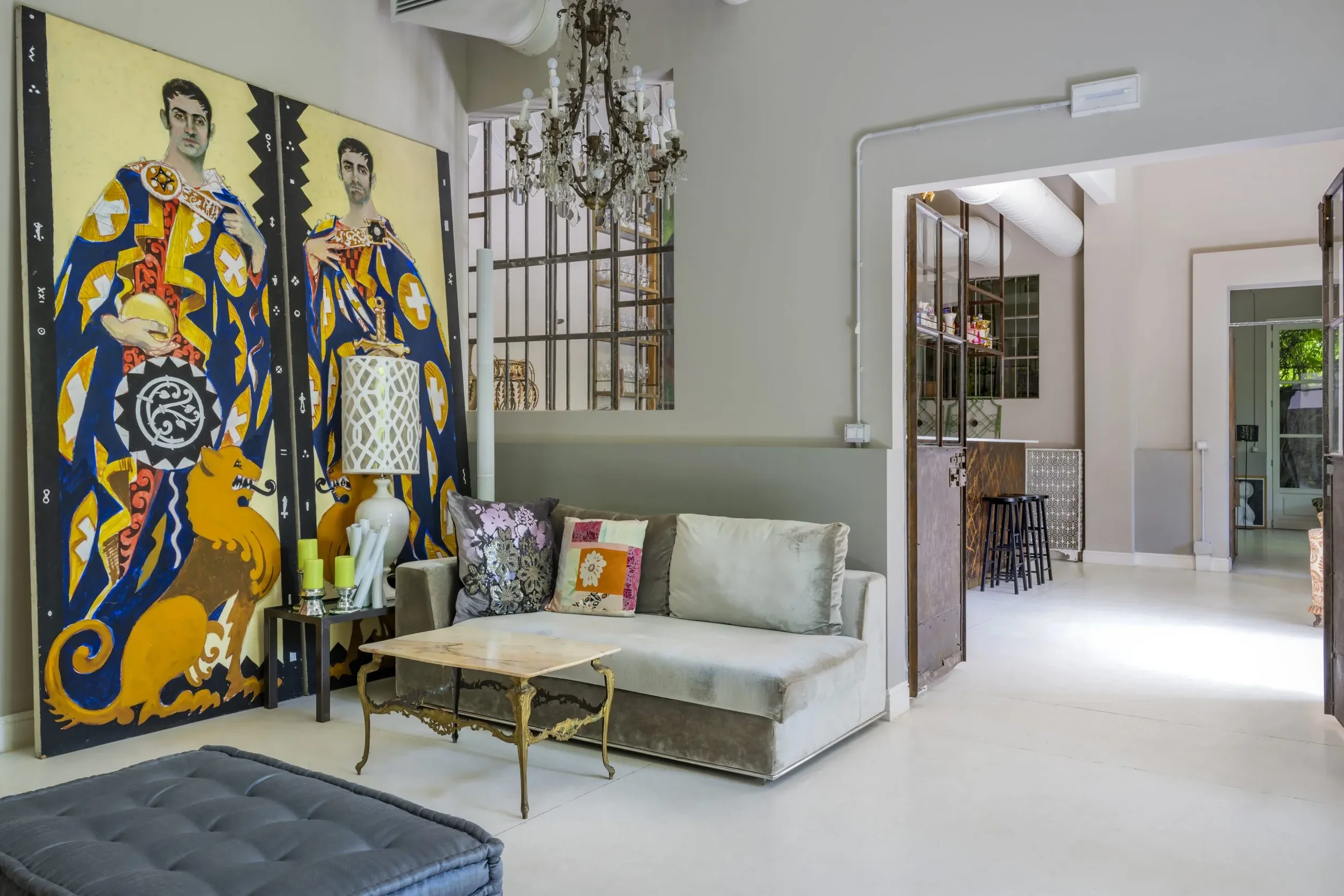 Interno di un soggiorno con parete decorata da due grandi dipinti con figure umane, divano con cuscini, sedia con lampada, e una porta aperta che porta a un'altra stanza. Mobili e decorazioni eleganti, lampadario di cristallo.
