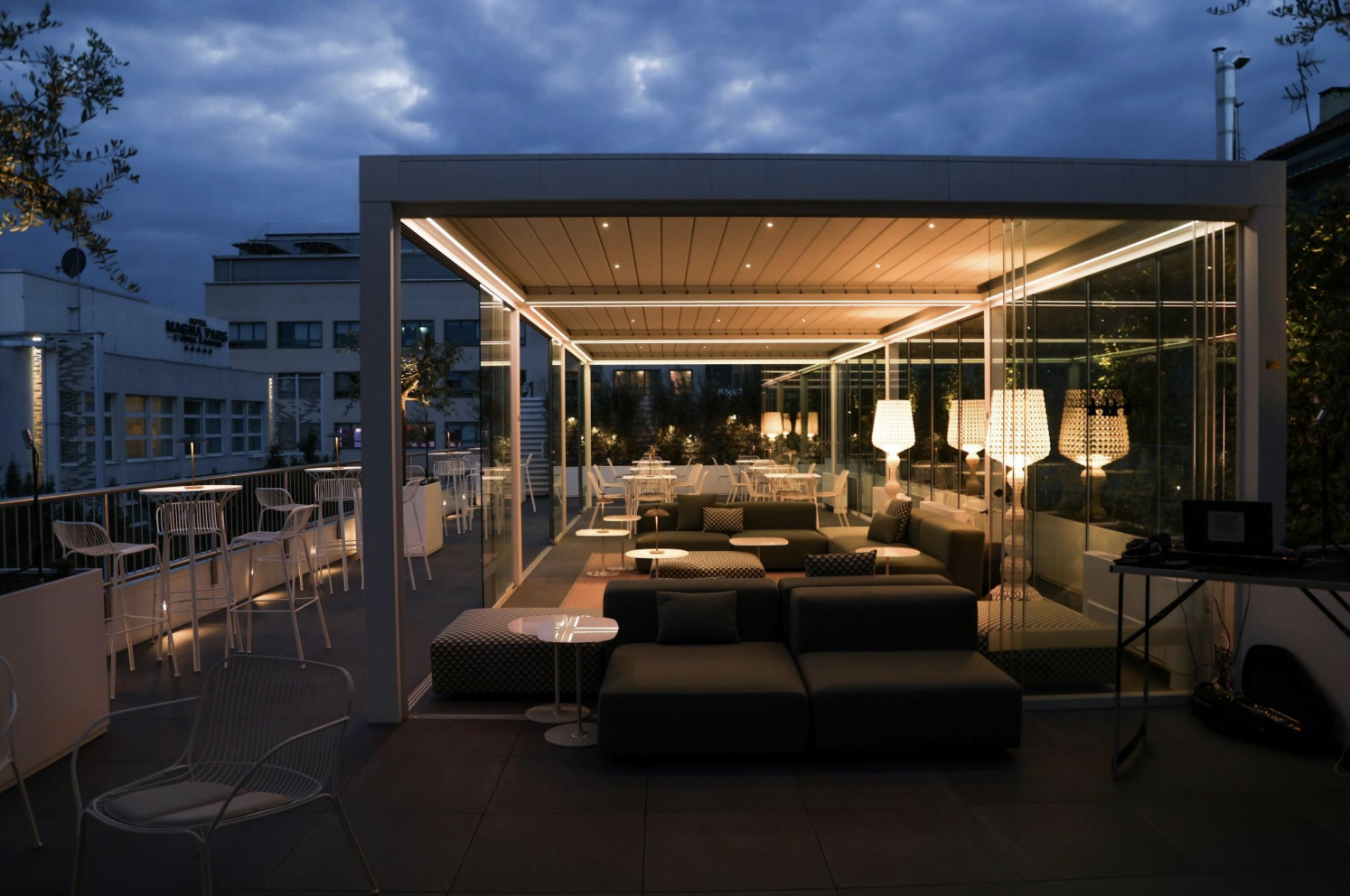 Terrazza con aree salotto e tavoli illuminati di sera, con luci soffuse e vista sulla città.