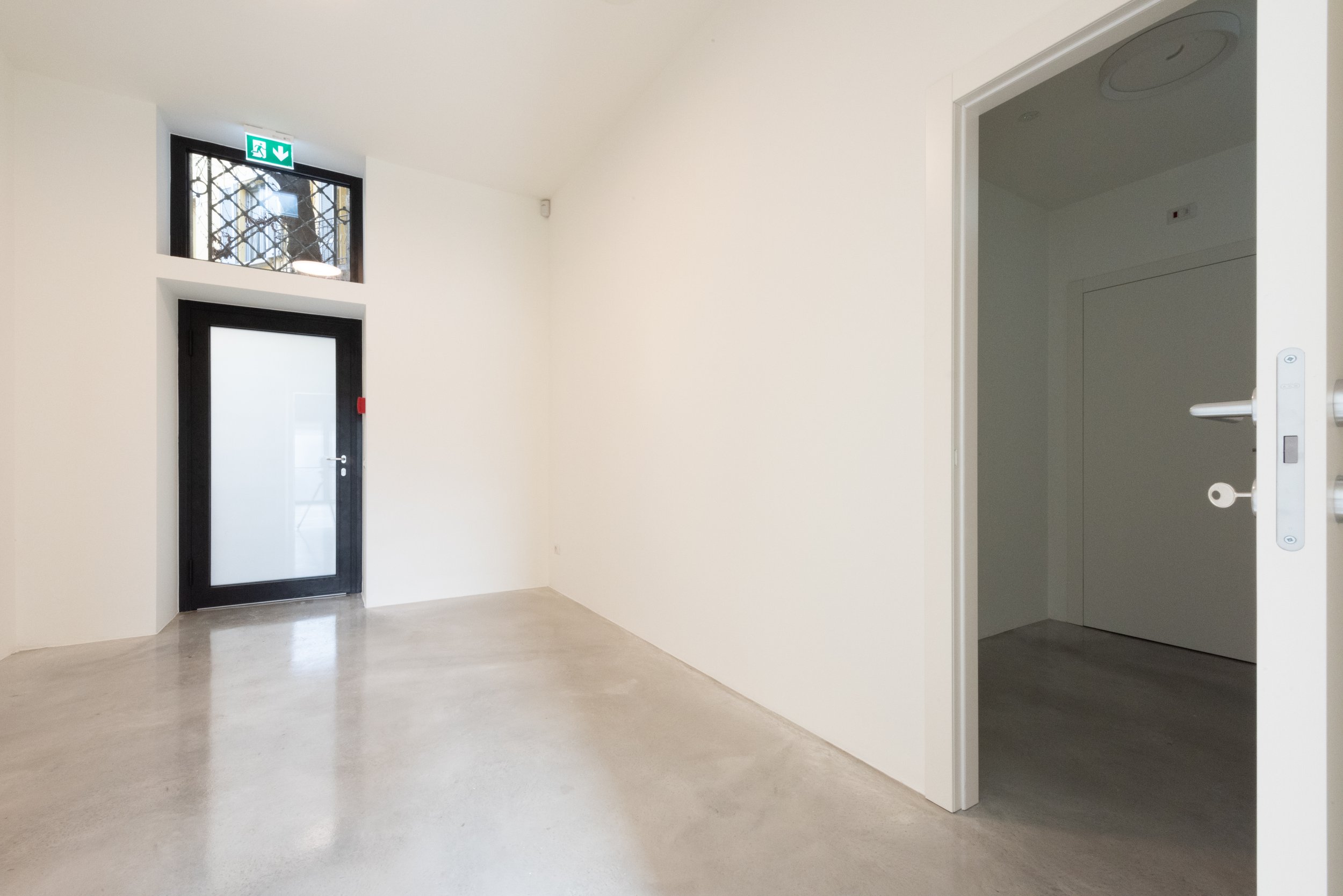 Stanza vuota con pareti bianche, porta con vetro e finestra sopra la porta, pavimento in cemento