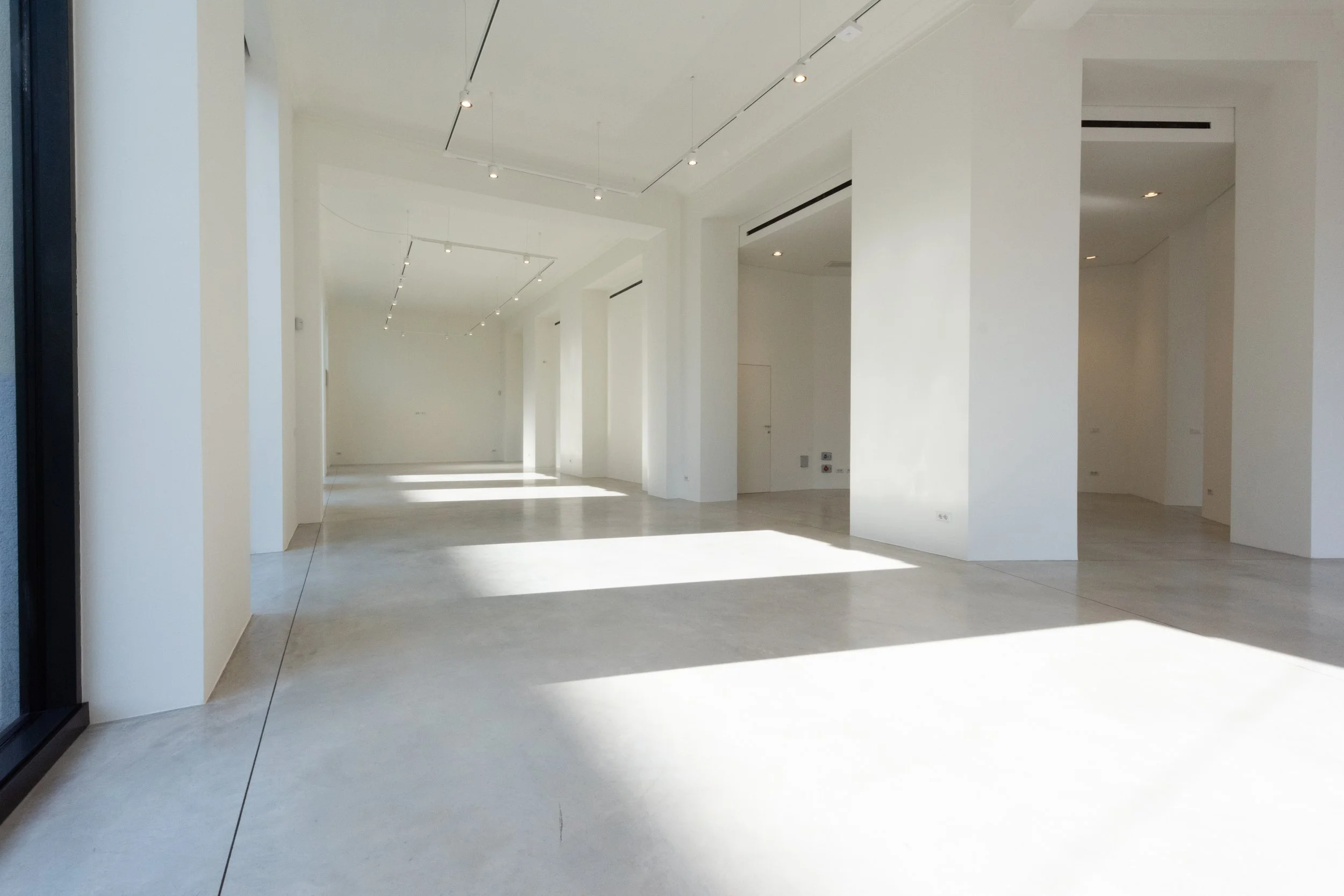Interno di uno spazio vuoto e moderno con pareti bianche, grande finestra e pavimento in cemento, illuminato dalla luce naturale.