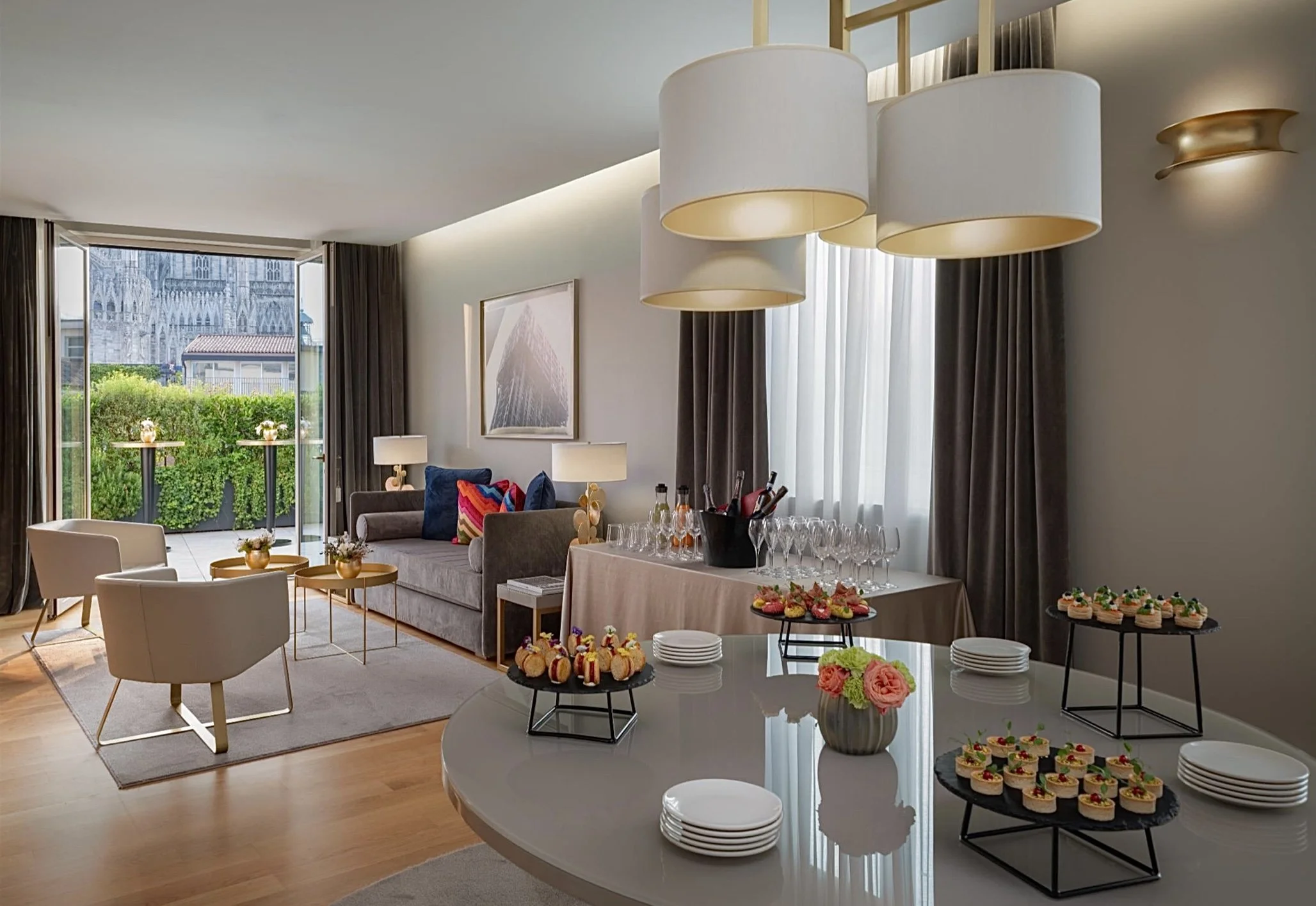 Interno di un soggiorno elegante con divano, poltrone e tavolini, balcone con vista al Duomo di Milano e tavolo con finger food e bevande