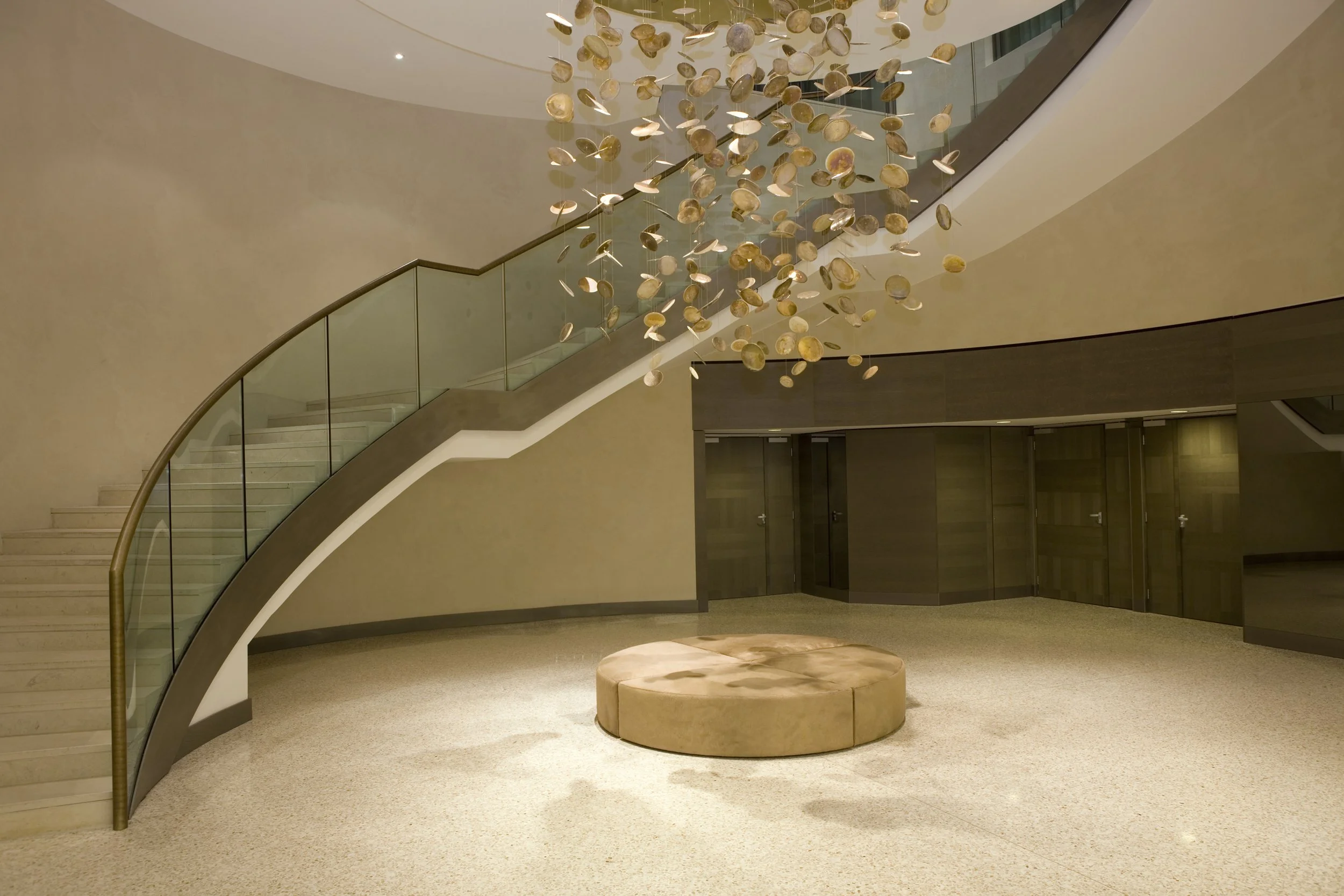 Interno di un edificio con scala a chiocciola in vetro e metallo, lampadario decorativo sospeso con piattini metallici, pareti beige, pavimento chiaro, panca rotonda al centro, porte in legno sullo sfondo.