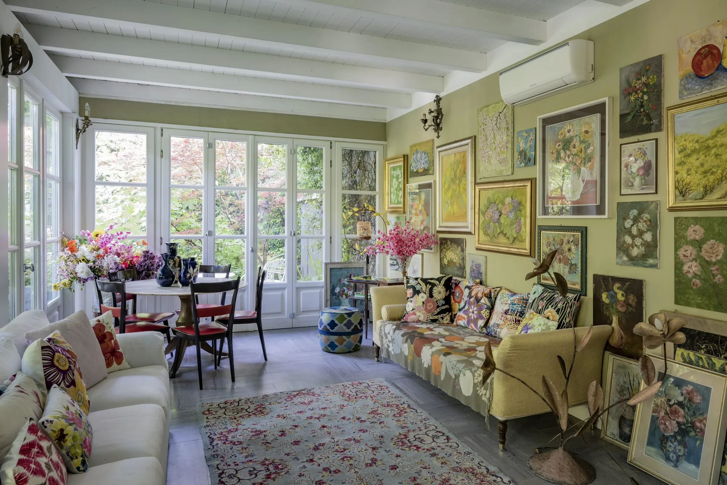 Soggiorno con parete decorata con molte opere d'arte di fiori, divani con cuscini colorati, tavolo rotondo con vasi di fiori, grande finestra con vista sul giardino esterno, pavimento di legno, tappeto dietro il divano.