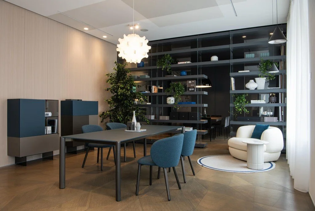 Interno di un soggiorno moderno con tavolo, sedie blu, scaffali neri con piante e oggetti decorativi, poltrona bianca, luci pendenti, e grande finestra con tende.