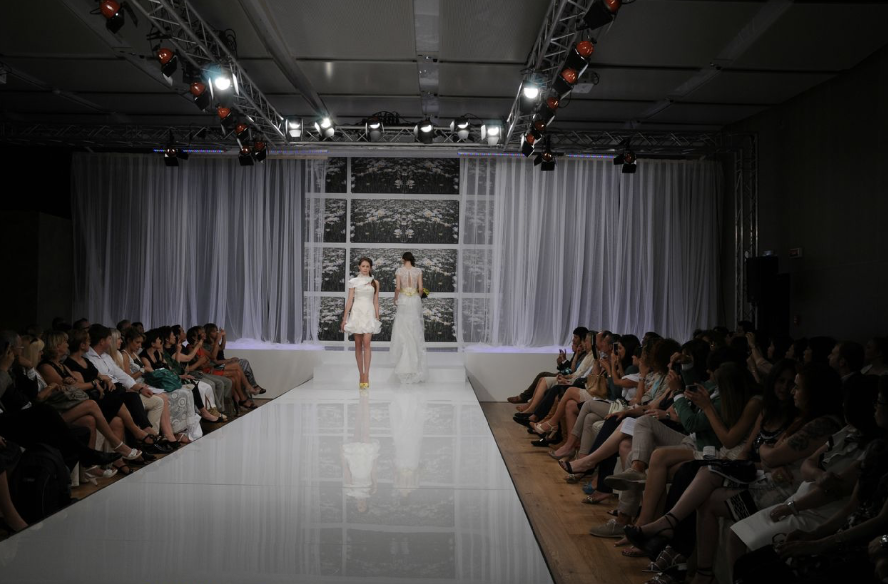 Modella cammina sulla passerella di una sfilata di moda, con pubblico seduto ai lati, luci e sfondo con tende bianche e finestra che mostra un paesaggio.