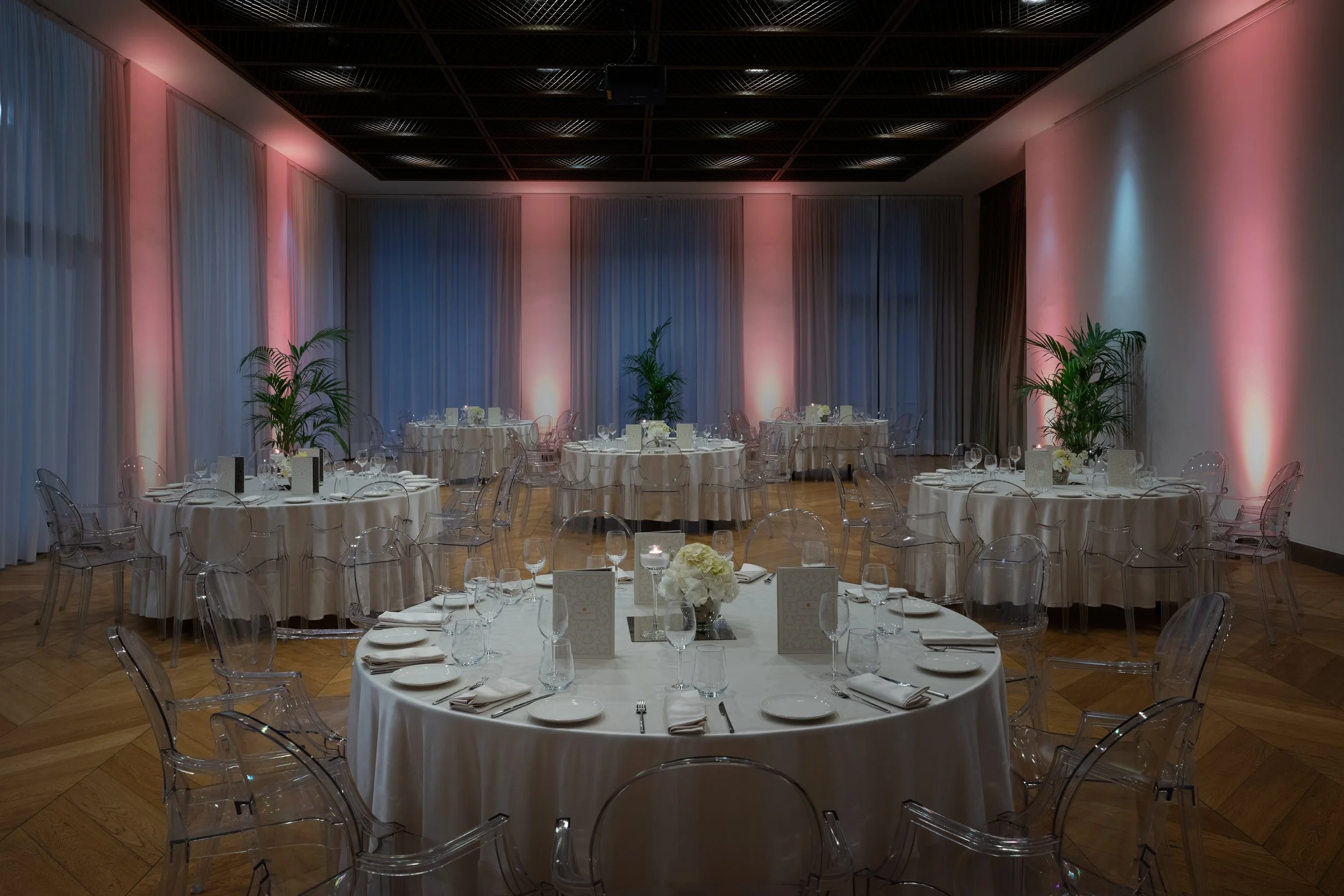 Sala elegantemente allestita con tavoli rotondi coperti da tovaglie bianche, decorati con composizioni floreali e set da cena, con luci soffuse e drappeggi alle pareti.