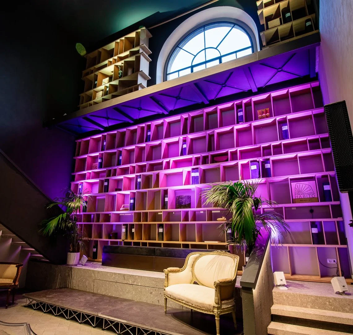 Una libreria colorata con scaffalature di legno illuminate da luci rosa e viola, con due piante verdi ai lati e una poltrona vintage al centro.