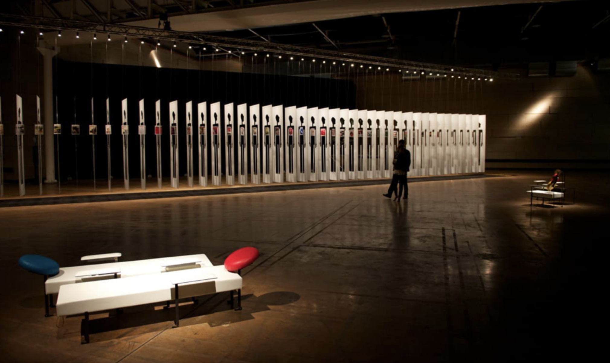 Installazione artistica in una galleria con numerosi strumenti musicali a forma di chitarra disposti in file, illuminata soffusamente, con sedie su un lato e due persone che osservano l'opera.