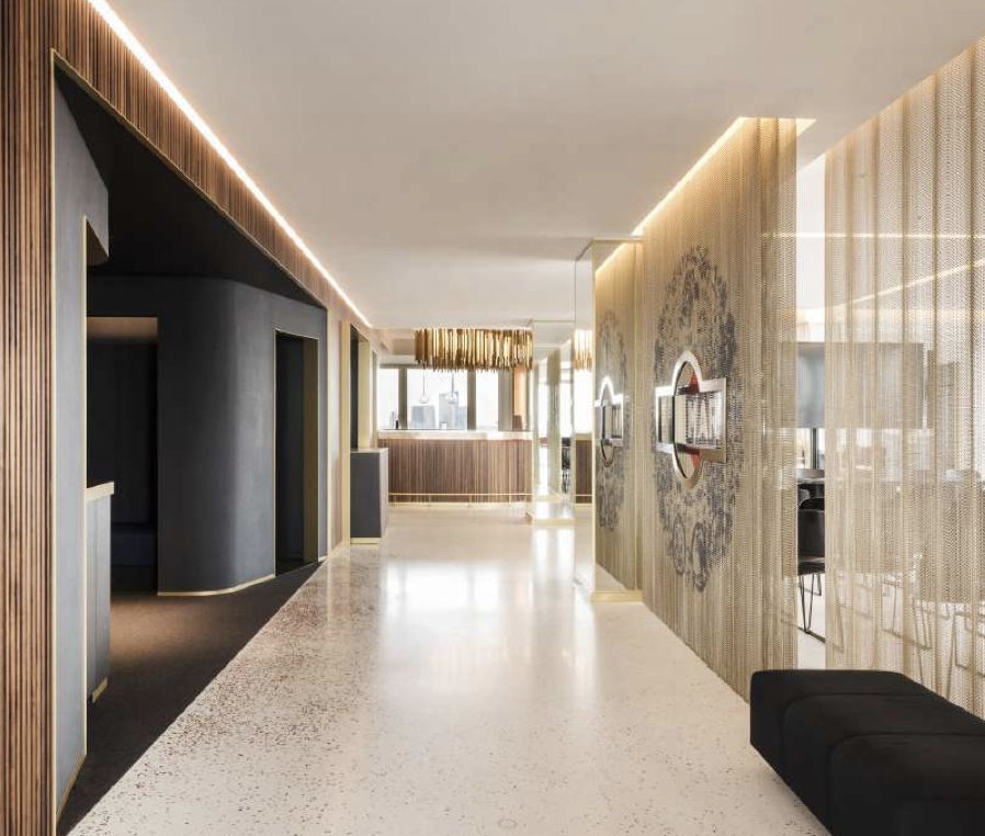 Interno di un hotel elegante con pavimento in marmo chiaro, pareti decorate con modanature e luci a LED, area reception sullo sfondo, parete divisoria di tessuto con decorazioni e schermi digitali, arredamento moderno e minimalista.