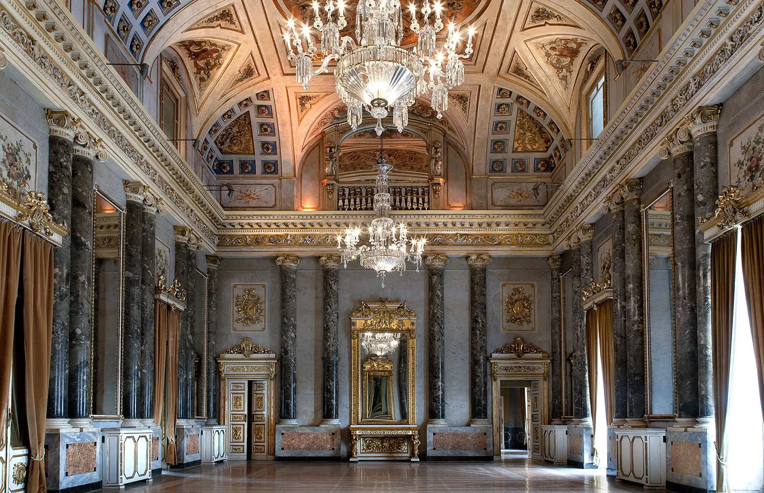 Sala da ballo con soffitto decorato, lampadari di cristallo e pareti ornate dorate e di marmo, con grandi finestre con tendaggi dorati.