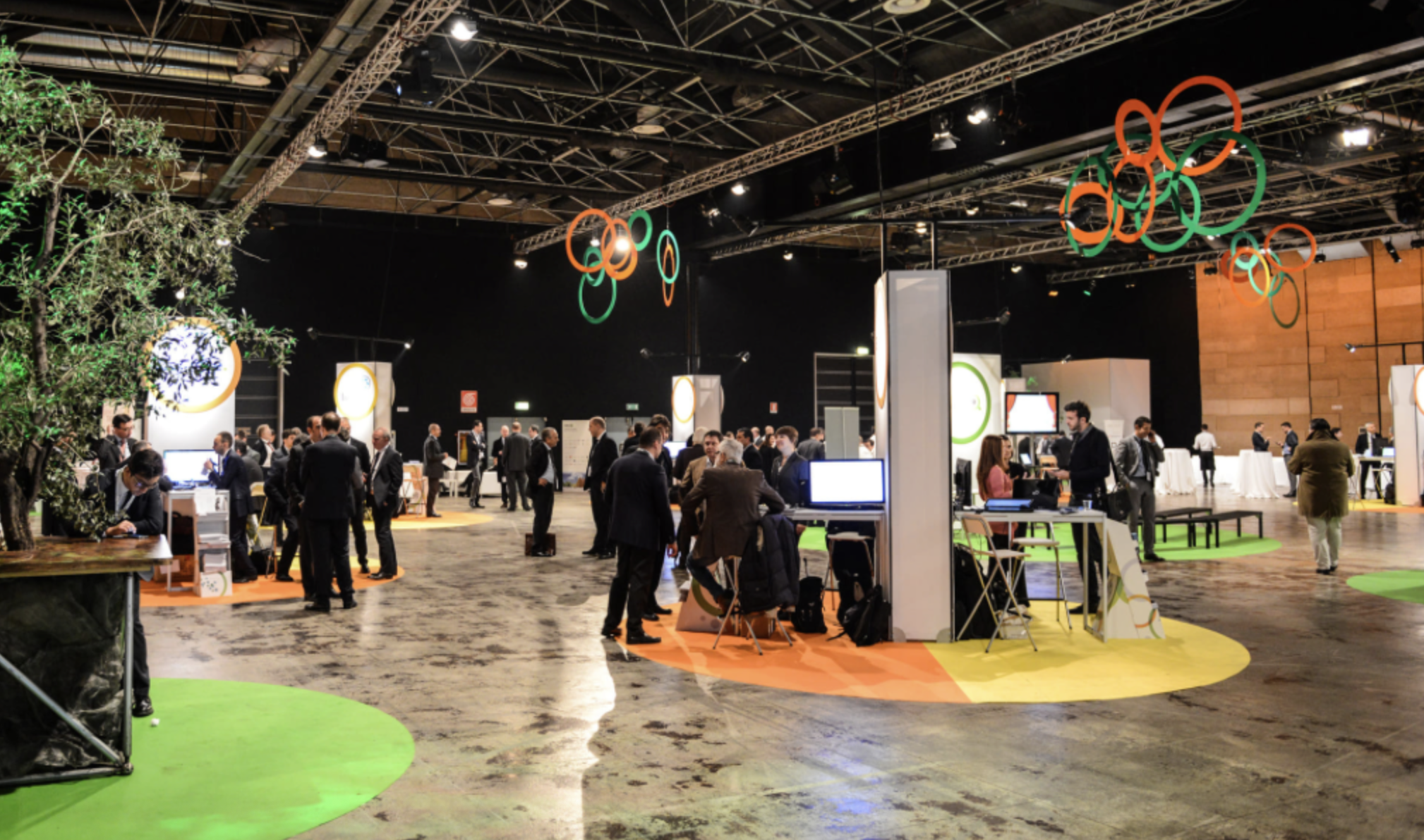 Una sala di conferenze affollata con persone che conversano e si spostano tra gli stand. Suspended circular decorazioni in arancio, verde e blu pendono dal soffitto, e ci sono aree con pavimentazione colorata in giallo, arancio e verde.