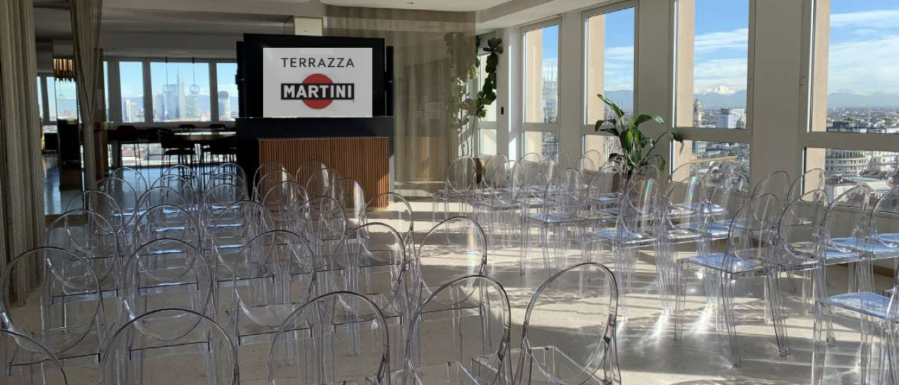 Sala con sedie di vetro trasparente e grandi finestre con vista sulla città, decorata con piante, e schermo con il logo 'Terrazza Martini'.