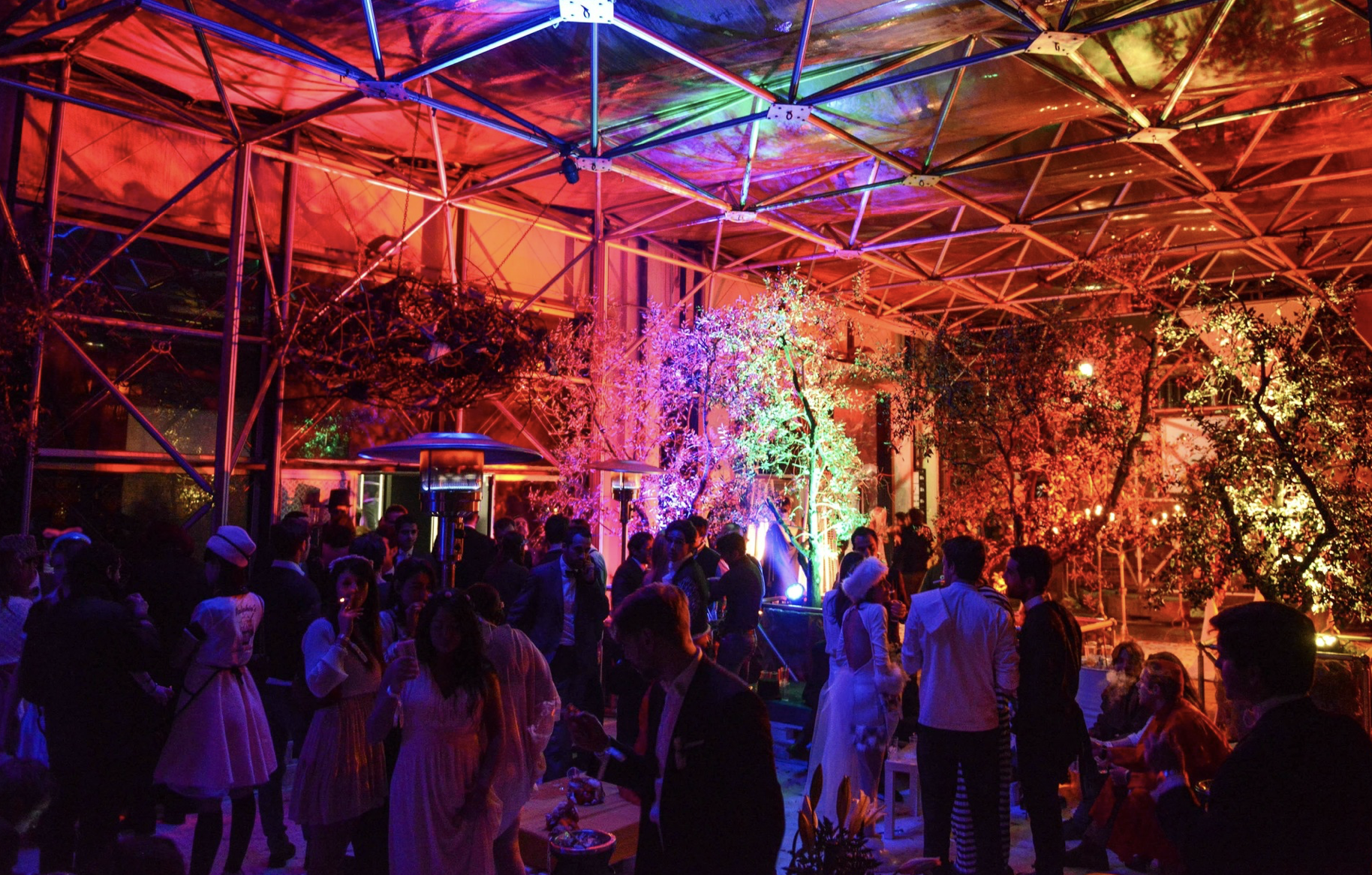 Una festa con molte persone sotto una tettoia moderna illuminata con luci colorate, decorazioni di alberi e luci luminose