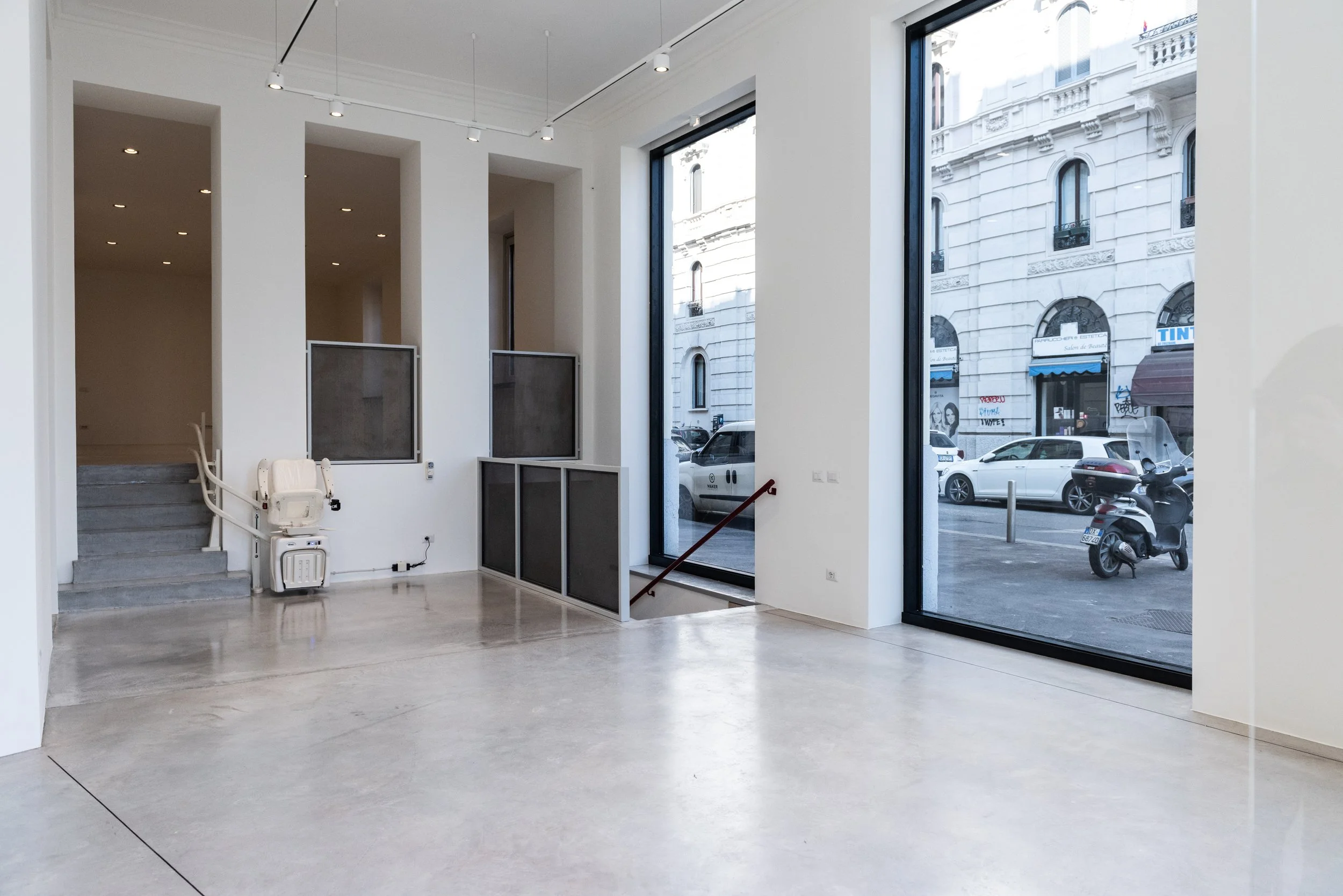 Interior di uno spazio commerciale o ufficio con grandi finestre che affacciano sulla strada, pareti bianche, pavimento in cemento, scala di pietra, e alcune strutture di supporto in metallo.