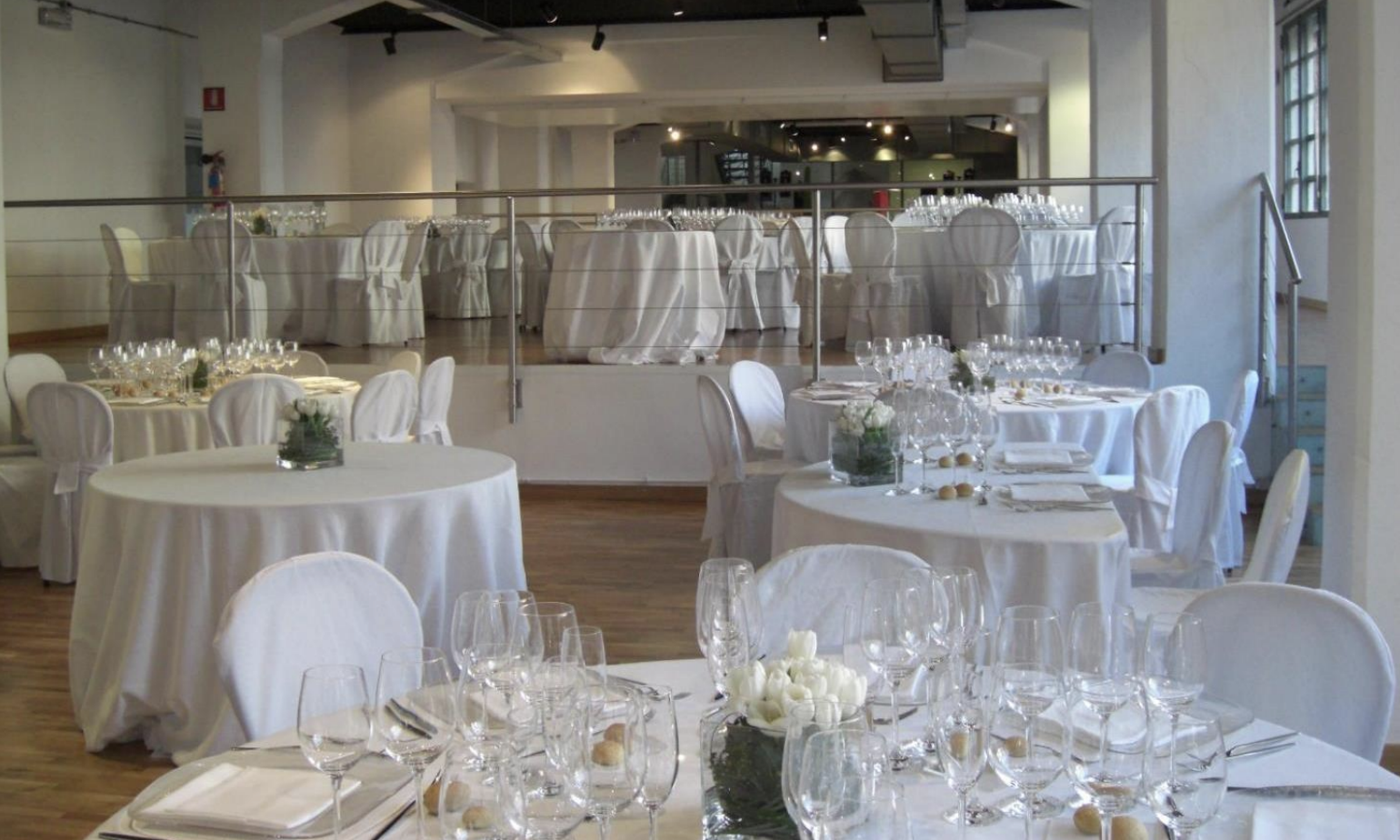 Sala per eventi o matrimonio con tavoli rotondi e quadrati decorati con tovaglie bianche, centrotavola di fiori bianchi, bicchieri da vino e piatti pronti per la cena. Sale luminose con grandi finestre e ambiente elegante.