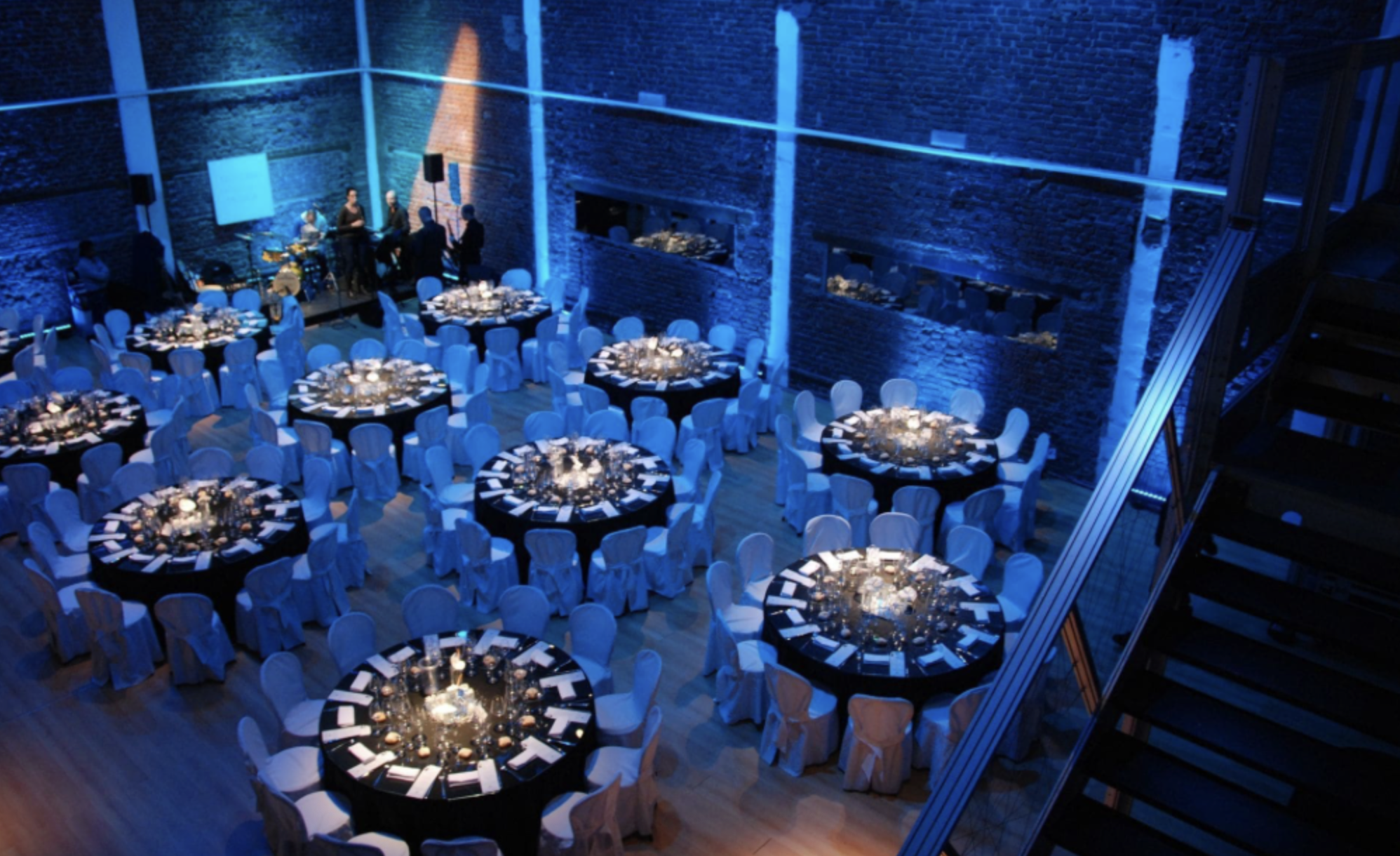 Sala elegante con tavoli rotondi vestiti con tovaglie nere, piatti, bicchieri e posate, illuminata da luci soffuse, con pareti di mattoni a vista e decorazioni luminose blu, disposto per un evento o ricevimento.