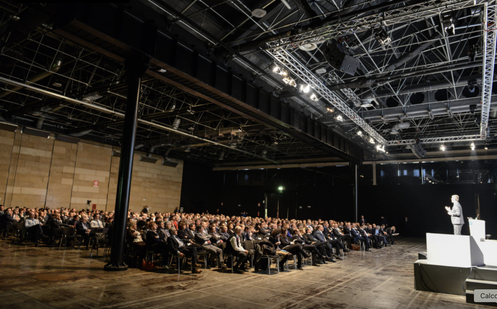 Pubblico seduto in un auditorium ascoltando un presentatore che parla sul palco