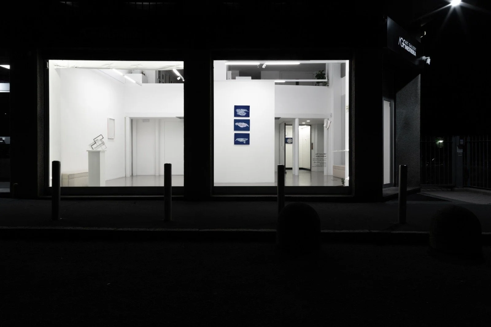 Interno di una galleria d'arte con pareti bianche, alcune opere d'arte e monitor, visibile attraverso vetrate illuminate di notte.