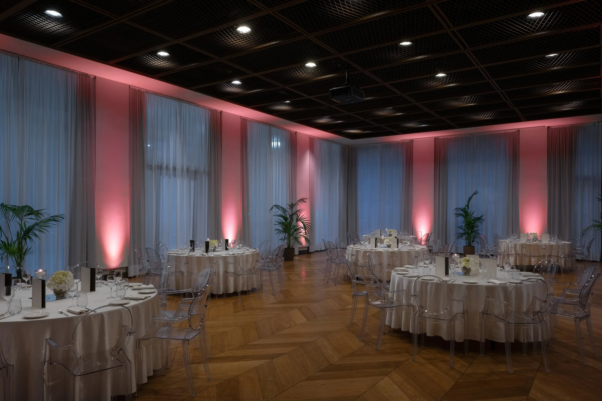 Sala elegante con tavoli tondi coperti da tovaglie bianche, decorati con fiori bianchi, bicchieri e posate, illuminata da luci rosa e tende bianche, con piante verdi sullo sfondo.