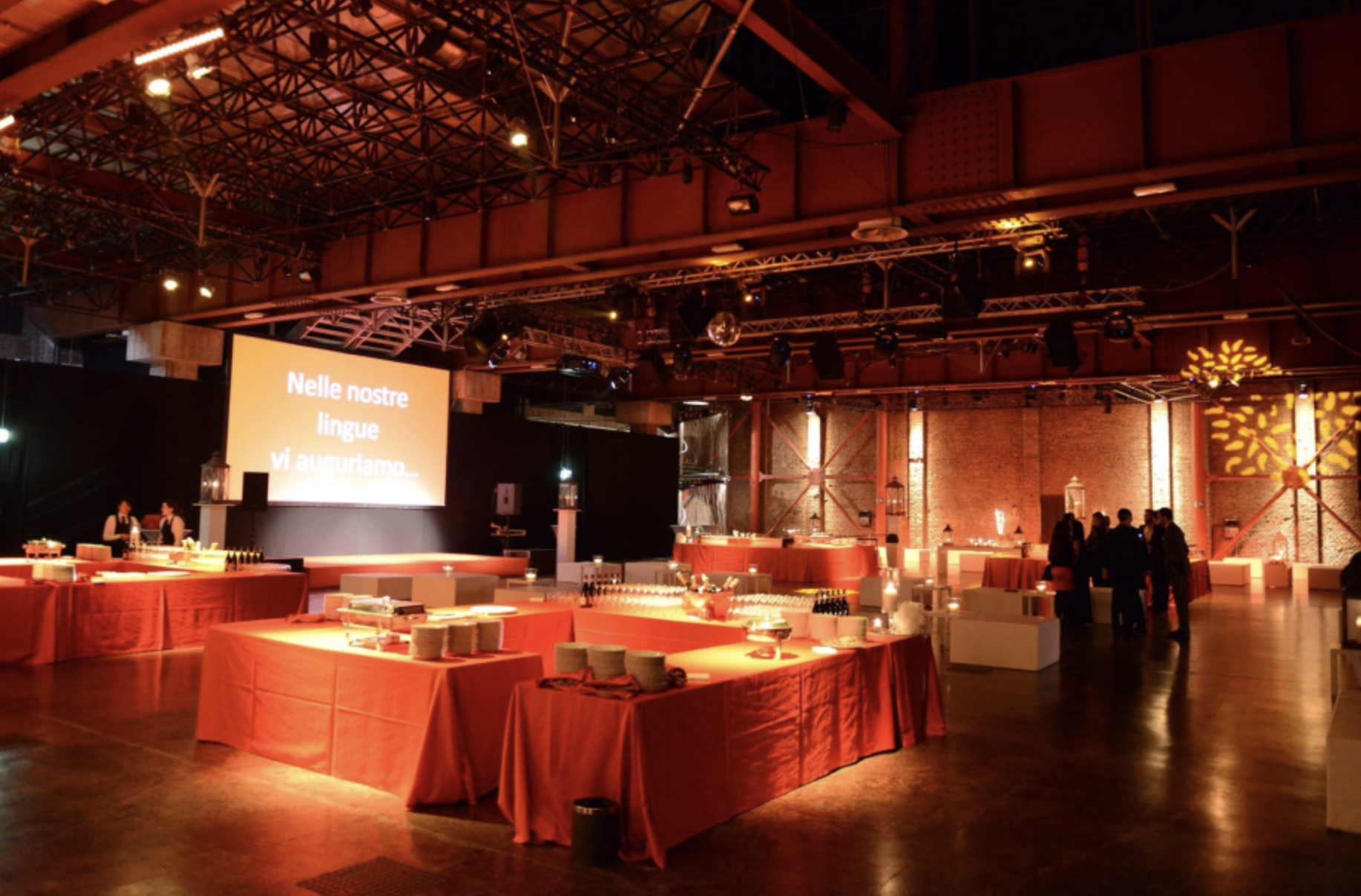 Sala elegante con tavoli coperti con tovaglie rosse, illuminazione calda, grande schermo con testo in italiano e alcuni gruppi di persone che conversano.