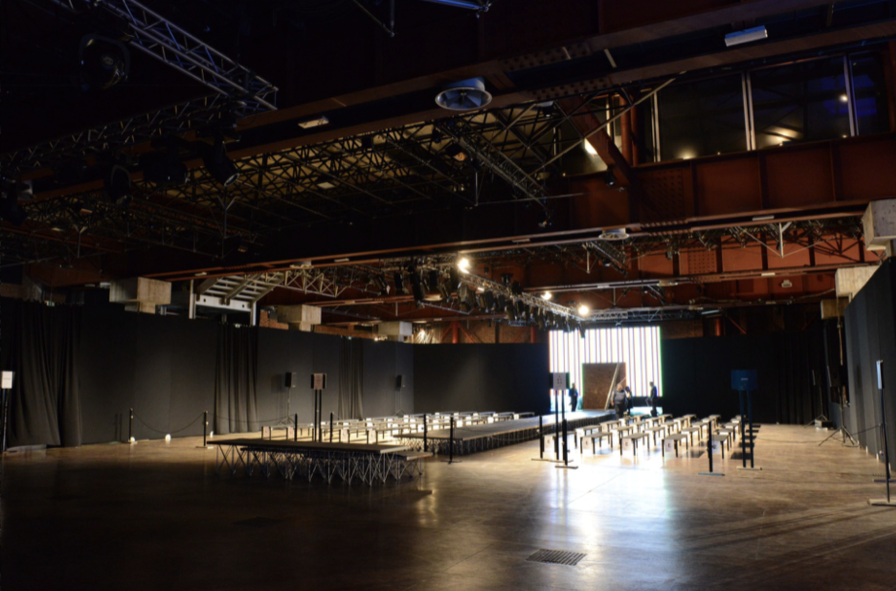 Interno di una sala di eventi o teatro, con luci ottiche e sedie disposte per uno spettacolo o conferenza, con alcune persone che si preparano dietro il palco.