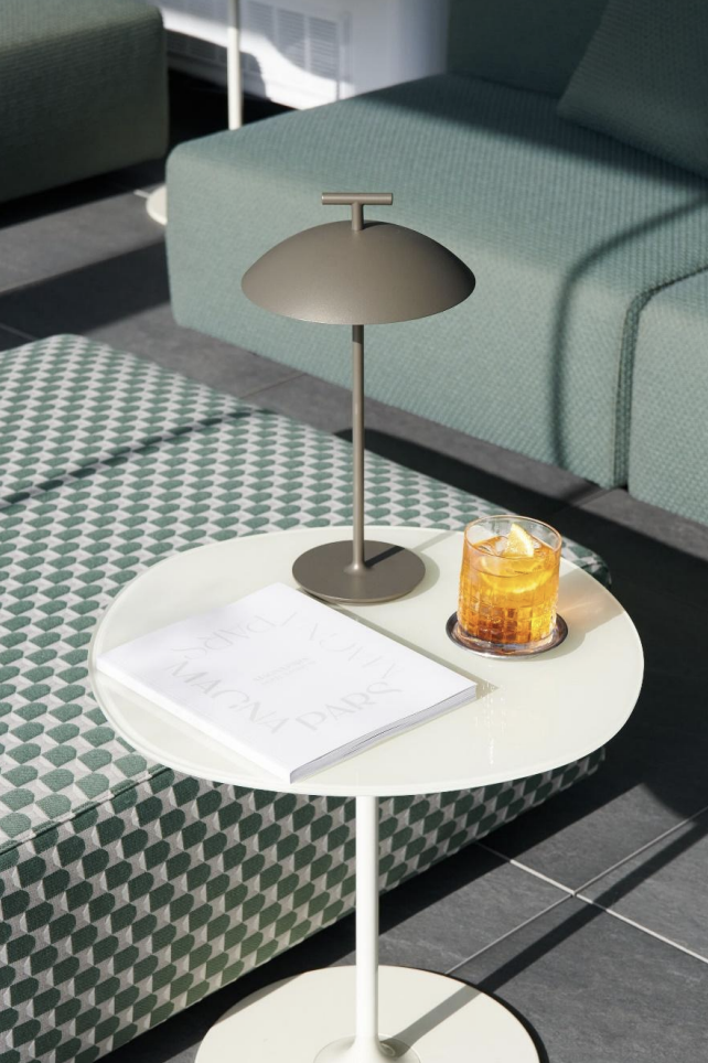 Tavolino con lampada da scrivania e un bicchiere con bevanda sull'ottomana. In sottofondo, divano verde e rivestimenti di mobili.