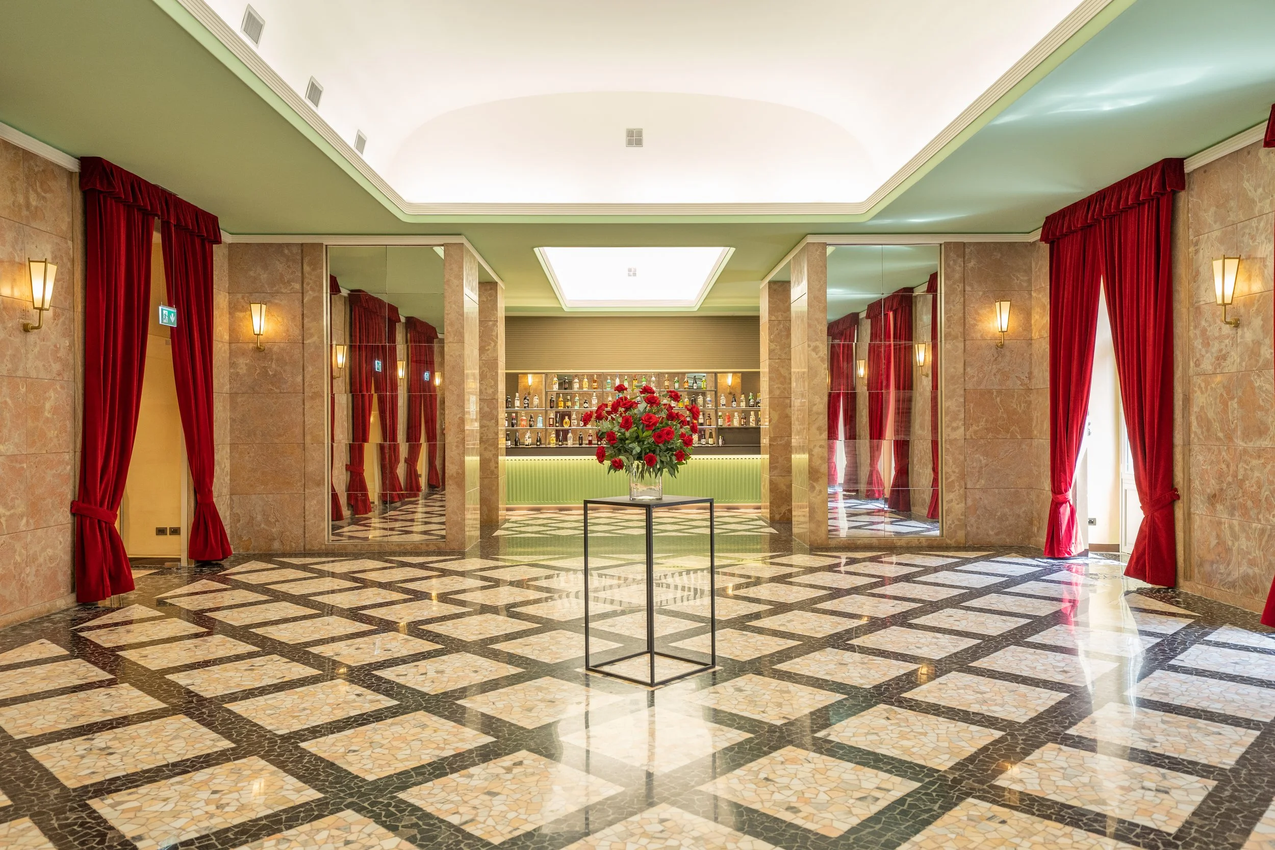 Ampio hall di hotel con pavimento in mosaico, pareti in marmo, grandi tende rosse, luci da parete e un bar sullo sfondo con scaffali pieni di bottiglie e un grande vaso con fiori rossi in centro.