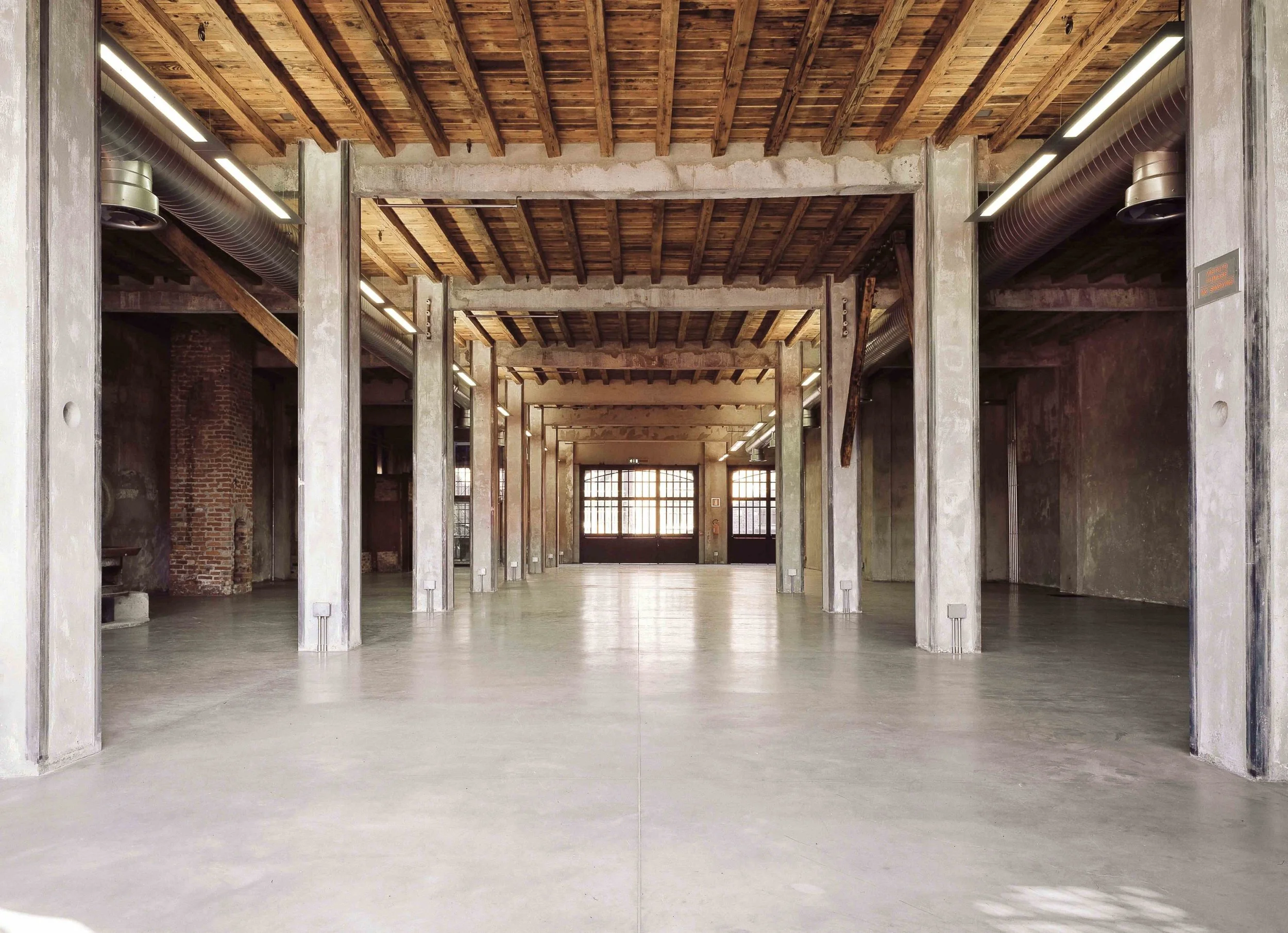 Interno di un edificio con colonne di cemento, soffitto in legno, grandi finestre e pavimento in cemento, atmosfera industriale.