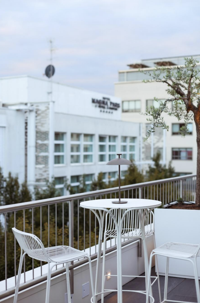 Balcone con tavolo e due sedie bianche, ombrellino e vista di edifici urbani e un albero lato destro.