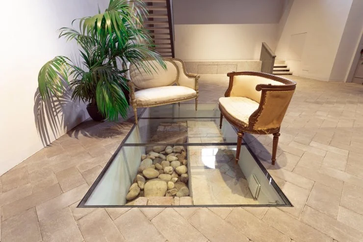 Due sedie di stile antico e una grande pianta in un ambiente interno con un pavimento di pietra e una zona con vetro e pietre sottostanti.