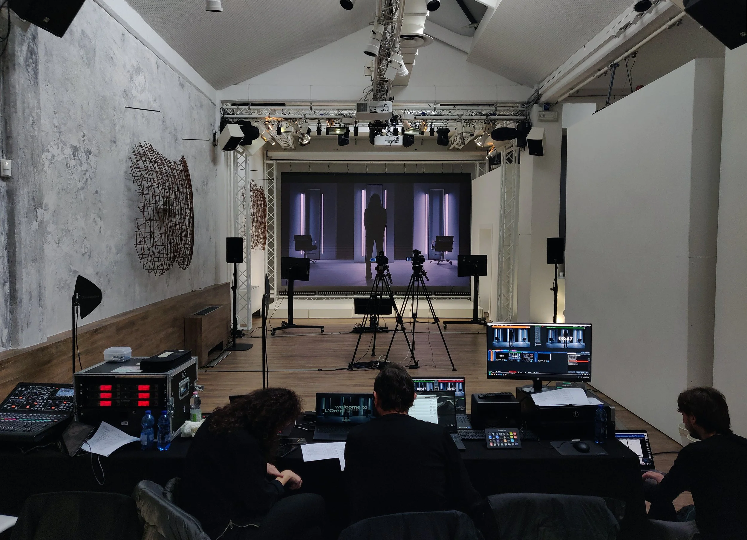 Una sala di registrazione con una grande scena in stile studio televisivo, attrezzatura per la produzione video, e tre persone sedute di fronte a monitor e computer, osservando la scena in scena
