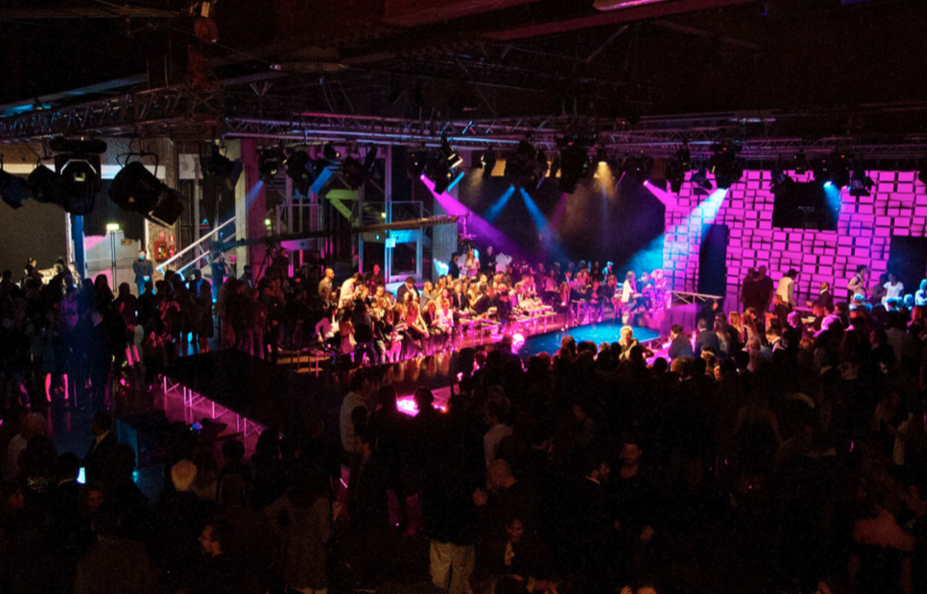 Interno di un teatro o sala concerti con molte persone sedute e in piedi, illuminata da luci colorate, con un palco centrale occupato da artisti e uno sfondo con pareti di luci