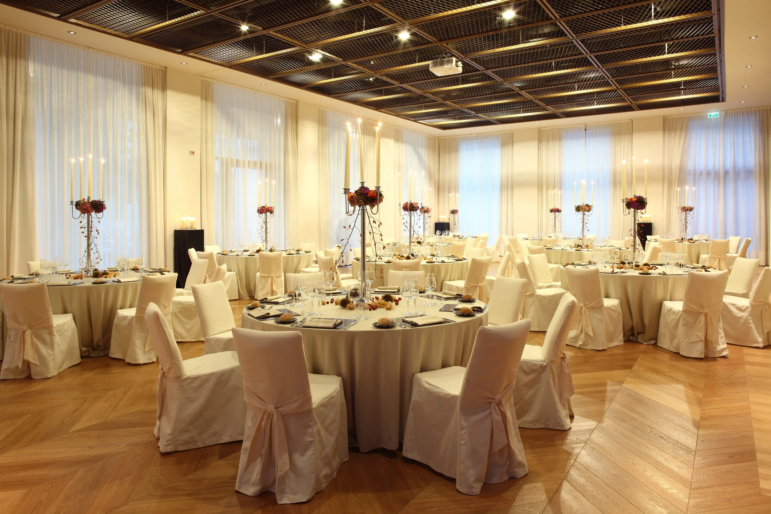 Salle da pranzo elegante con tavoli rotondi coperti con tovaglie beige, sedie con coperture bianche e decorazioni floreali centrali con candelabri e candele. Pareti con grandi tende bianche e soffitto in legno a griglia.