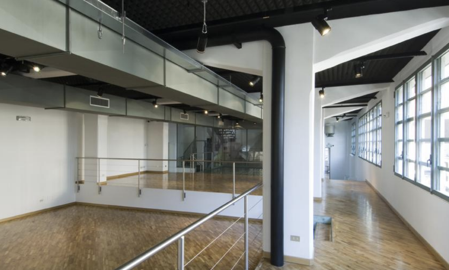 Interno di un edificio con pareti bianche, finestre grandi, pavimento in legno chiaro, strutture in metallo nero e soffitto nero con luci a spot.
