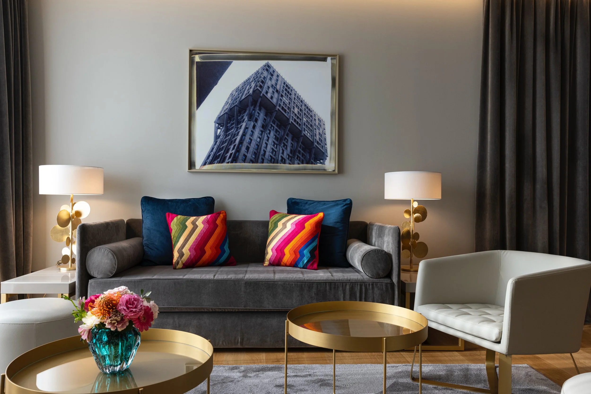 Interno di un soggiorno elegante con divano grigio, due poltrone bianche, tavolini dorati, lampade da tavolo, cuscini colorati con zigzag, un quadro di un edificio moderno e fiori in un vaso di vetro