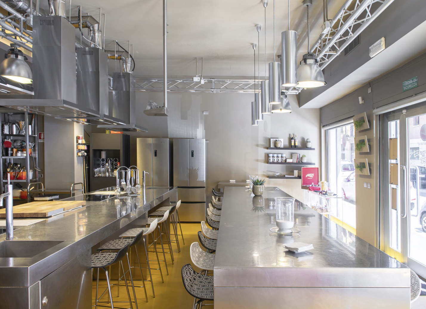 Interno di un bar o ristorante moderno con bancone in acciaio, sgabelli, frigoriferi, scaffali con spezie e oggetti decorativi, finestre grandi che lasciano entrare molta luce naturale e alcune piante.